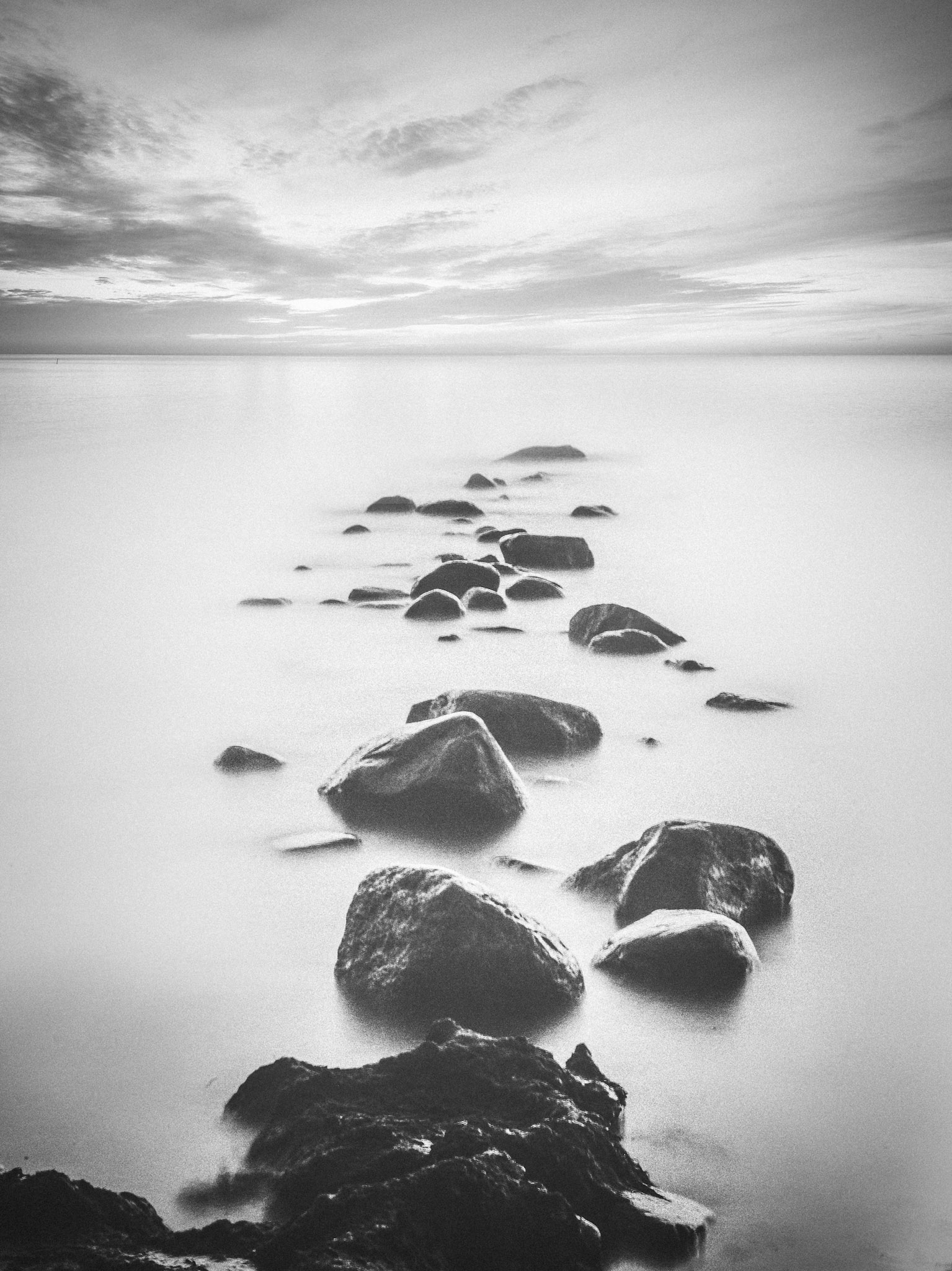 Svartvitt fotografi av stenar som leder ut i stilla vatten.