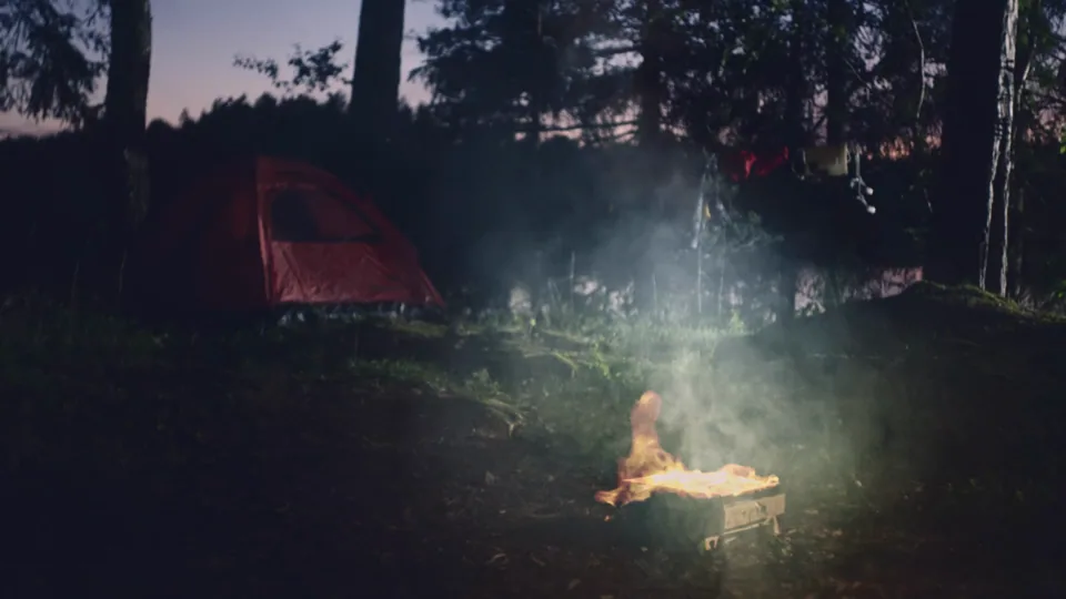Filmstill: ett tält och en lägereld i skogen i skymningen.