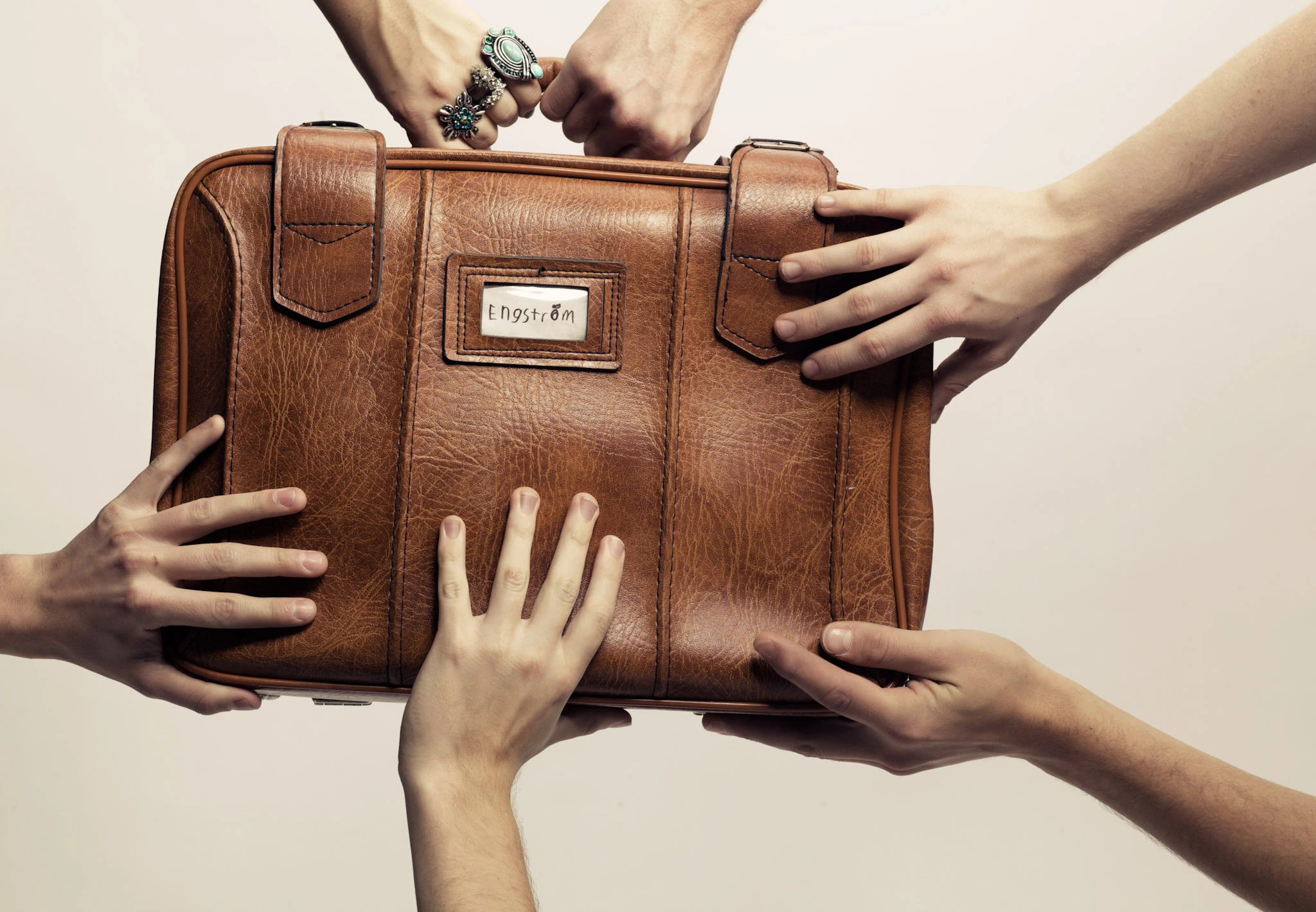 Several hands reach around a brown vintage suitcase against a pale background.