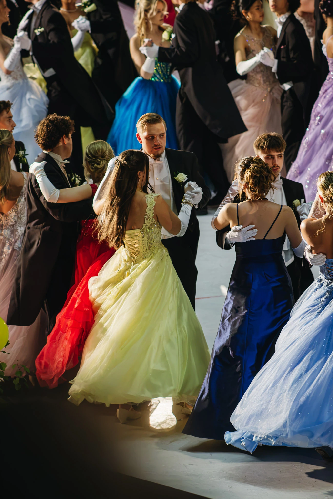 Par dansar i långa gula, blå och röda klänningar under riktat ljus.