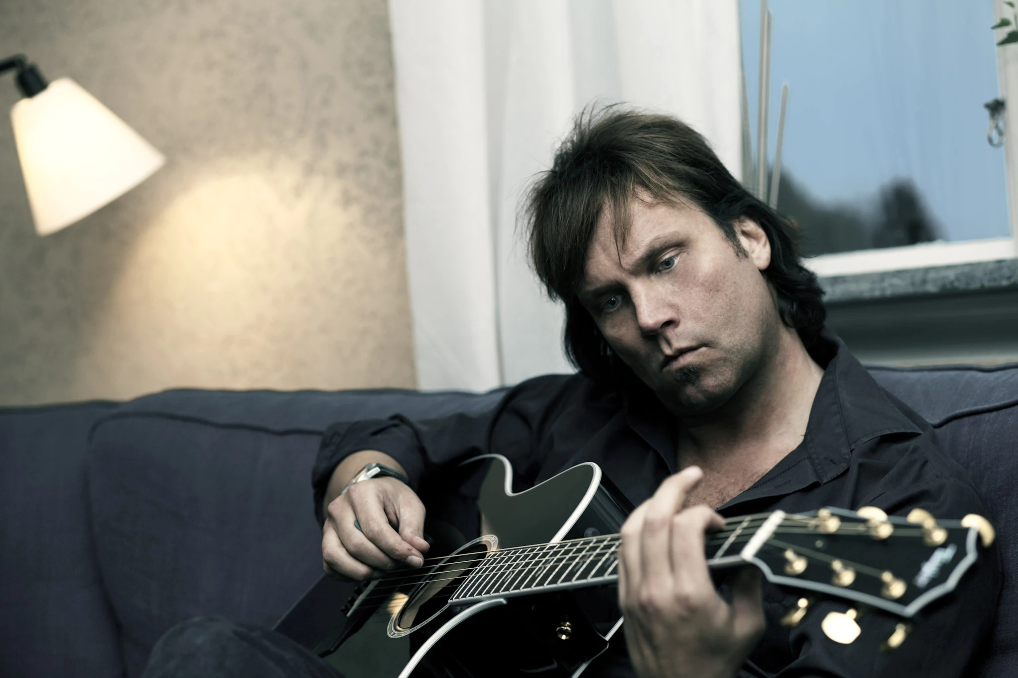 En man med akustisk gitarr sitter i en mörk soffa under en tänd läslampa.