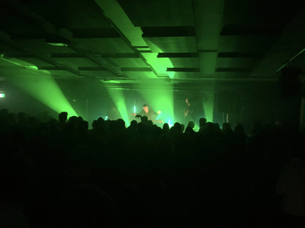 Audience silhouettes in front of green stage light beneath large square acoustic panels.