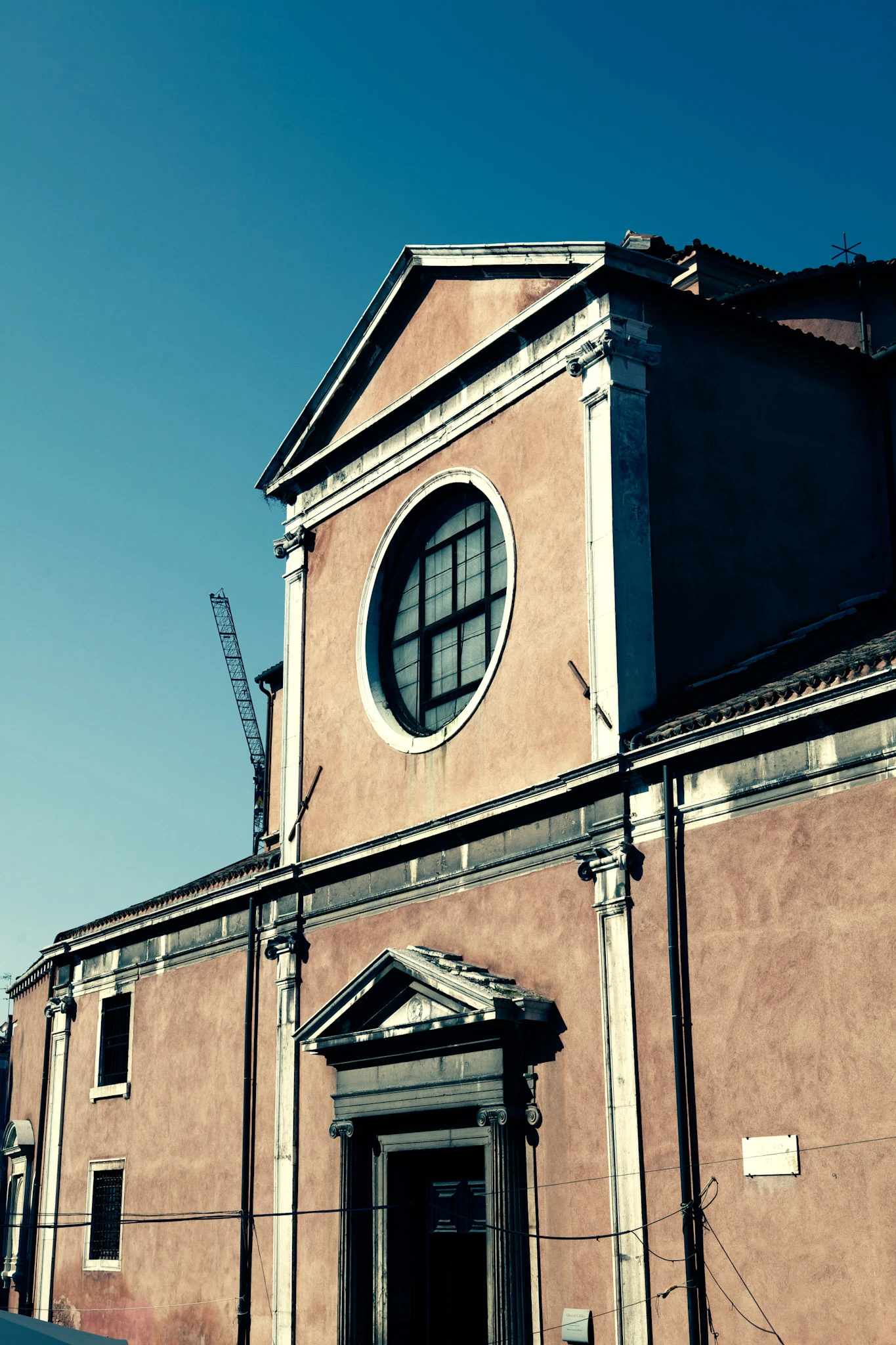 A pink plastered facade with a large round window, gable and dark door against a turquoise sky.