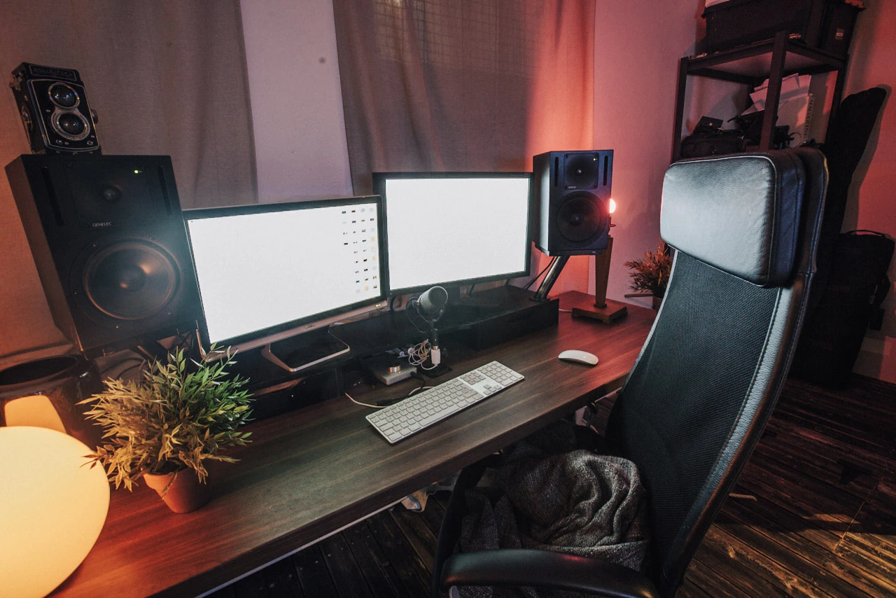 Two monitors and large speakers in front of an office chair in red and warm lamplight.
