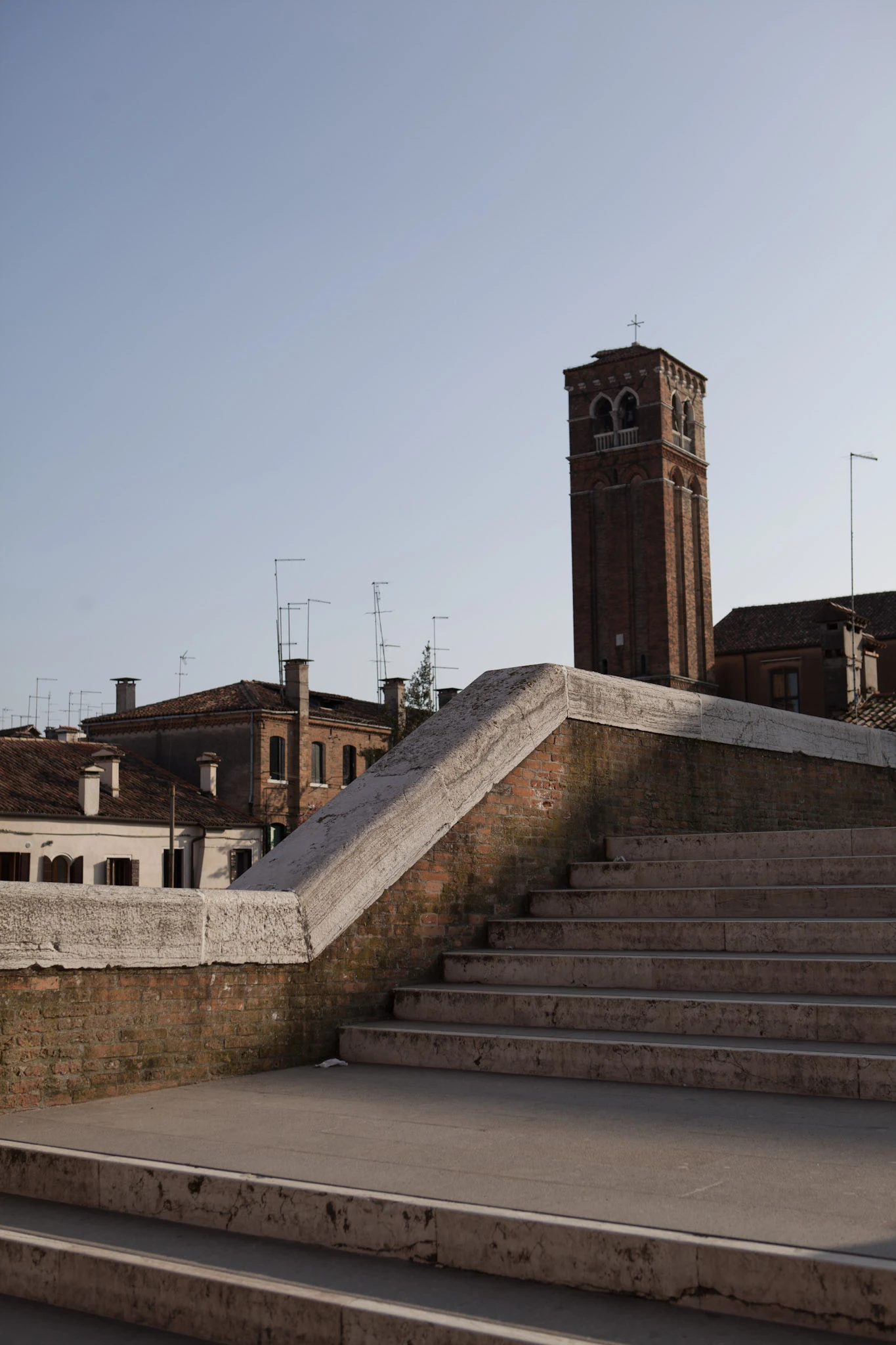 Broad stone steps lead towards low rooftops and a tall brick tower beneath a pale sky.