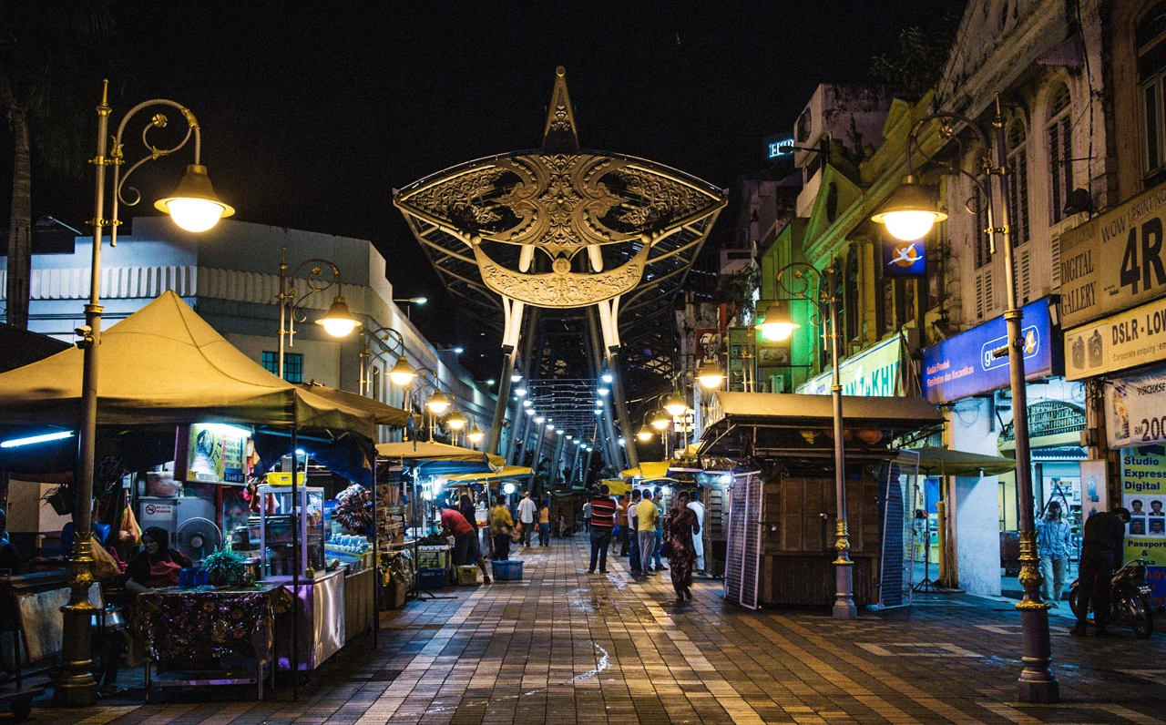 A lit city street with market stalls, people and a large decoration overhead.