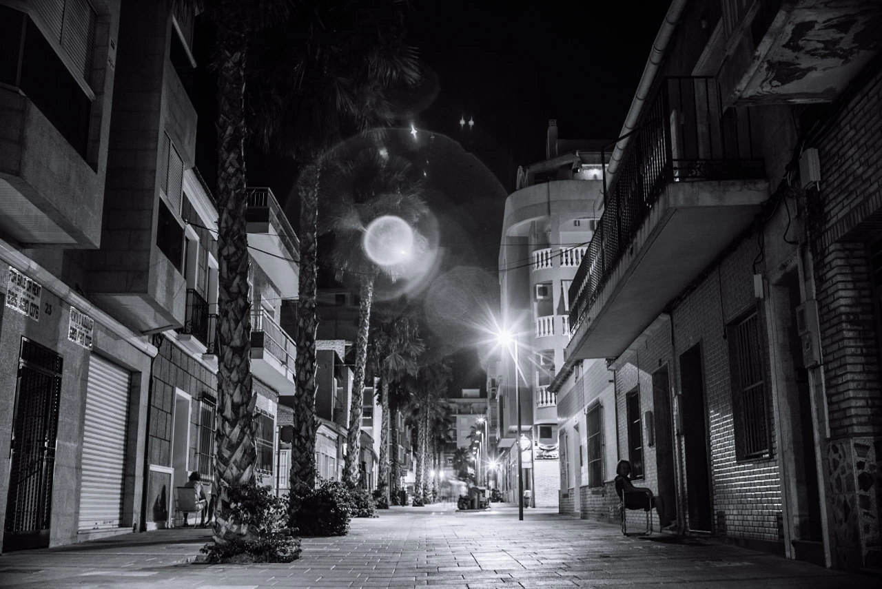 An empty city street with streetlights between tall façades in black and white.