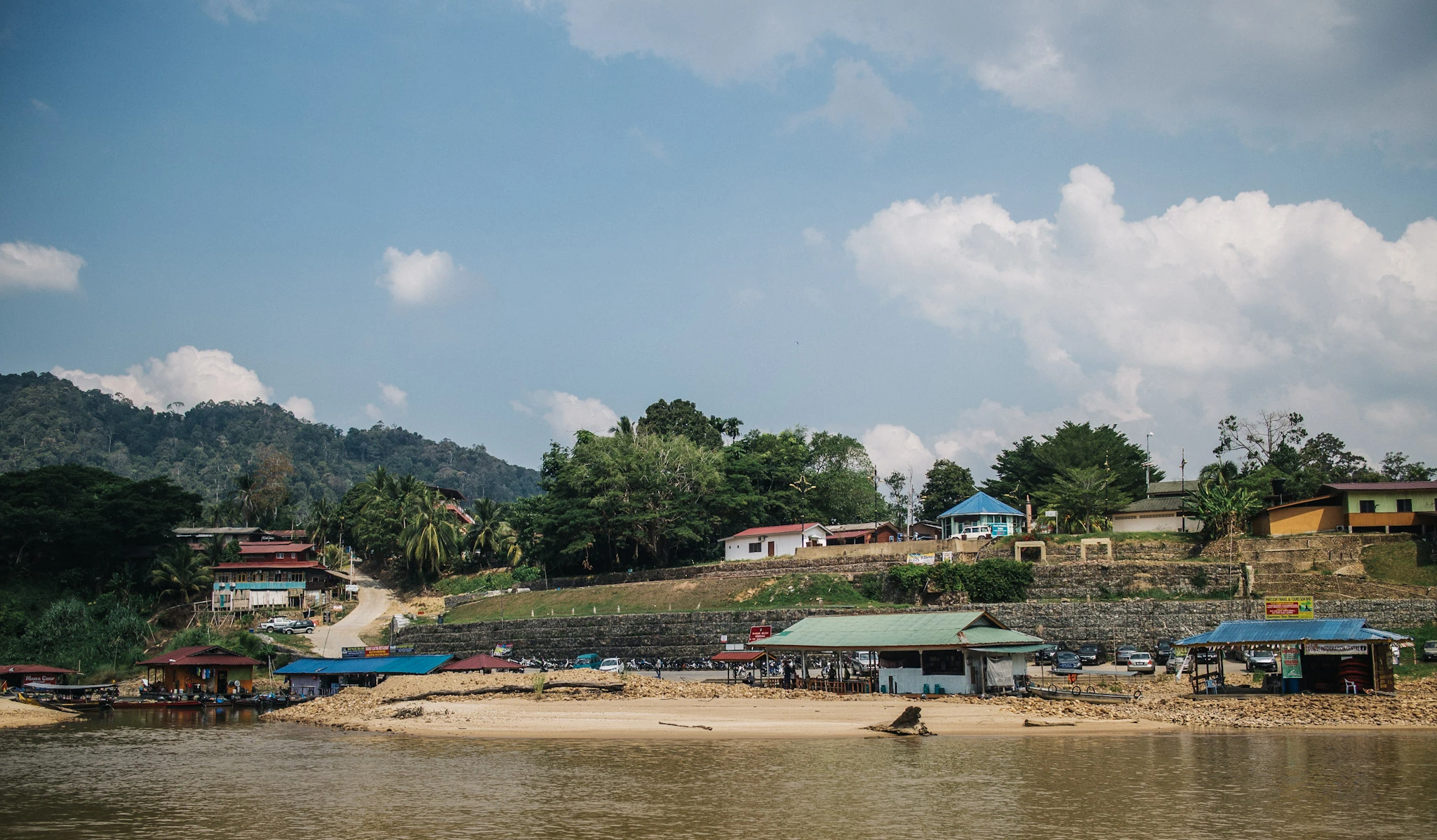 Low buildings sit between a brown river and a wooded hill.