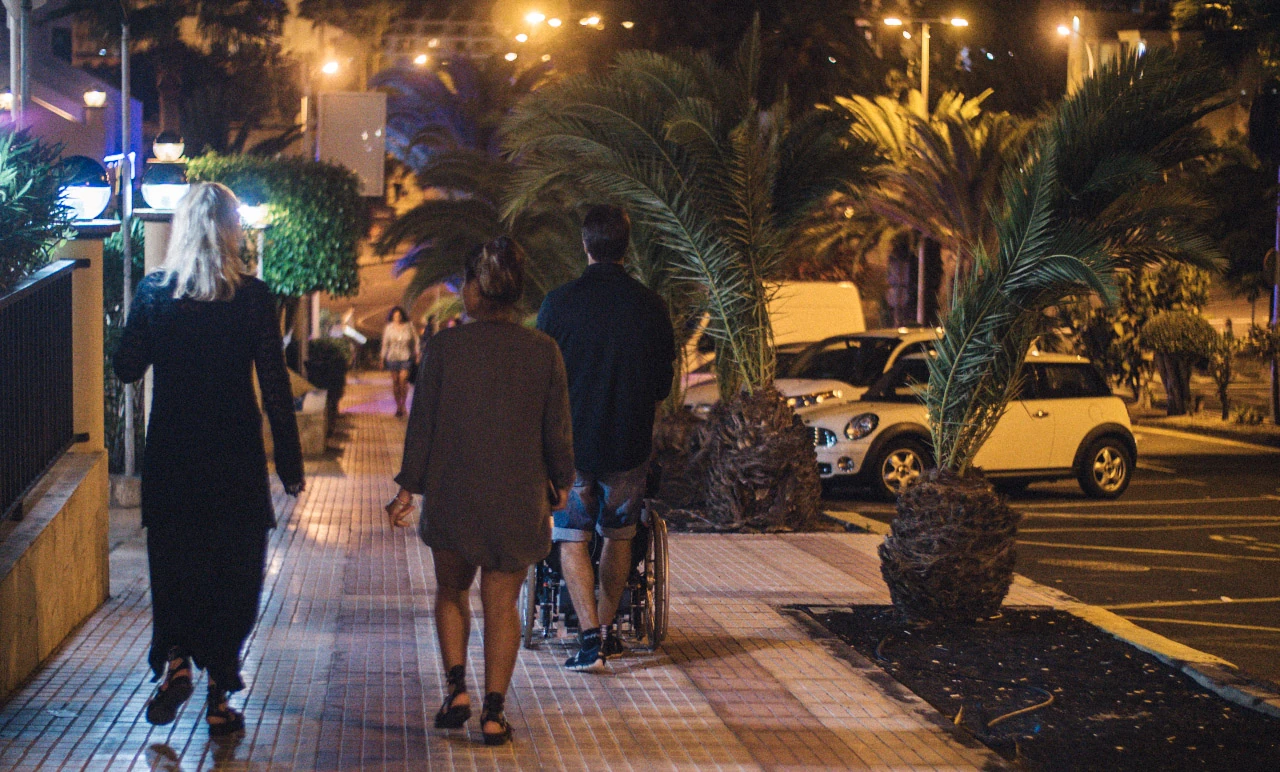 People walk along a palm-lined pavement under warm streetlights at night.
