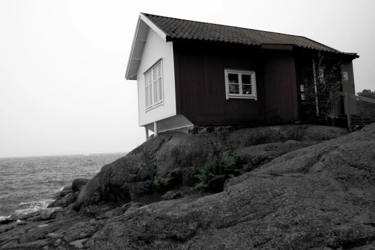 Ett litet hus på en klippkant över havet i svartvitt.