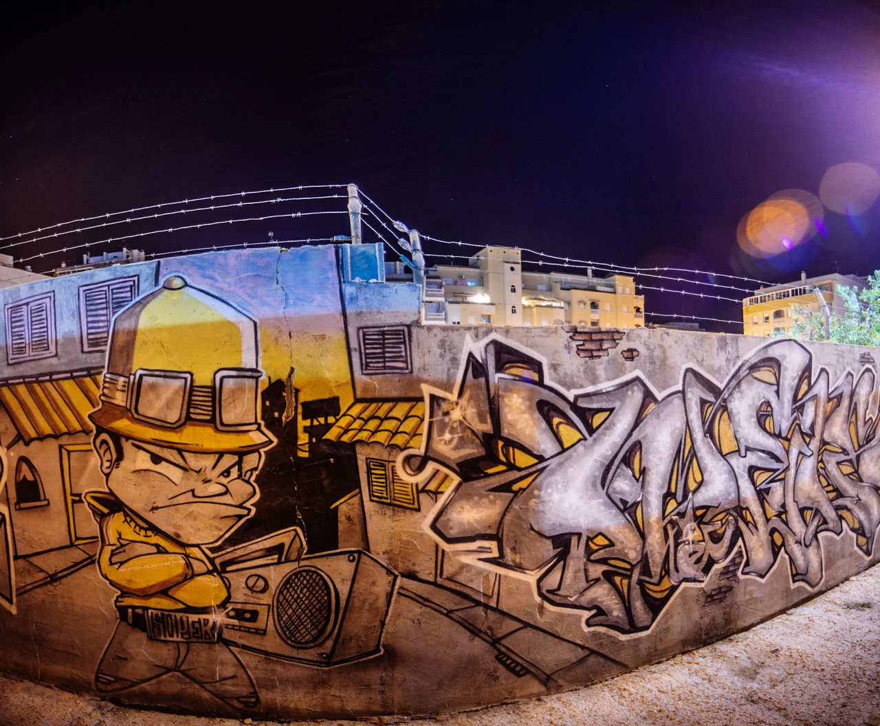 A graffiti-covered wall at an illuminated street corner beneath a dark sky.