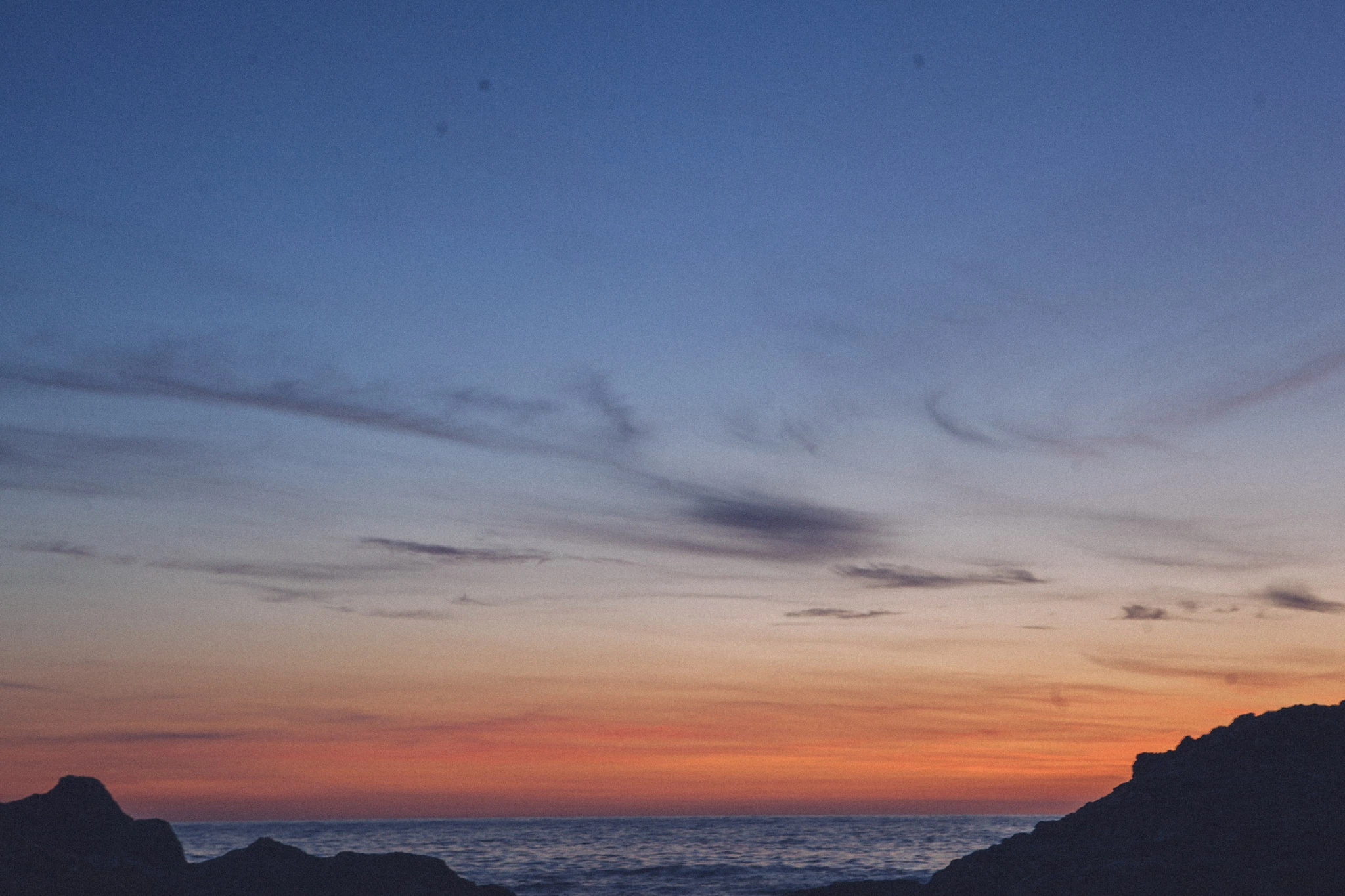Dark coastal silhouettes beneath thin clouds and an orange horizon after sunset.