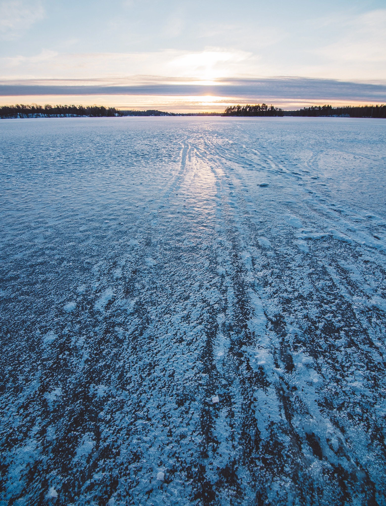 A frozen lake with long marks in the ice beneath a pale blue and pink evening sky.