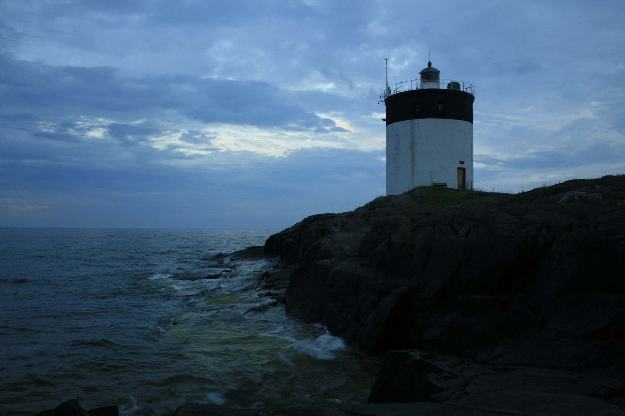 A lighthouse on dark rocks above blue water at dusk.