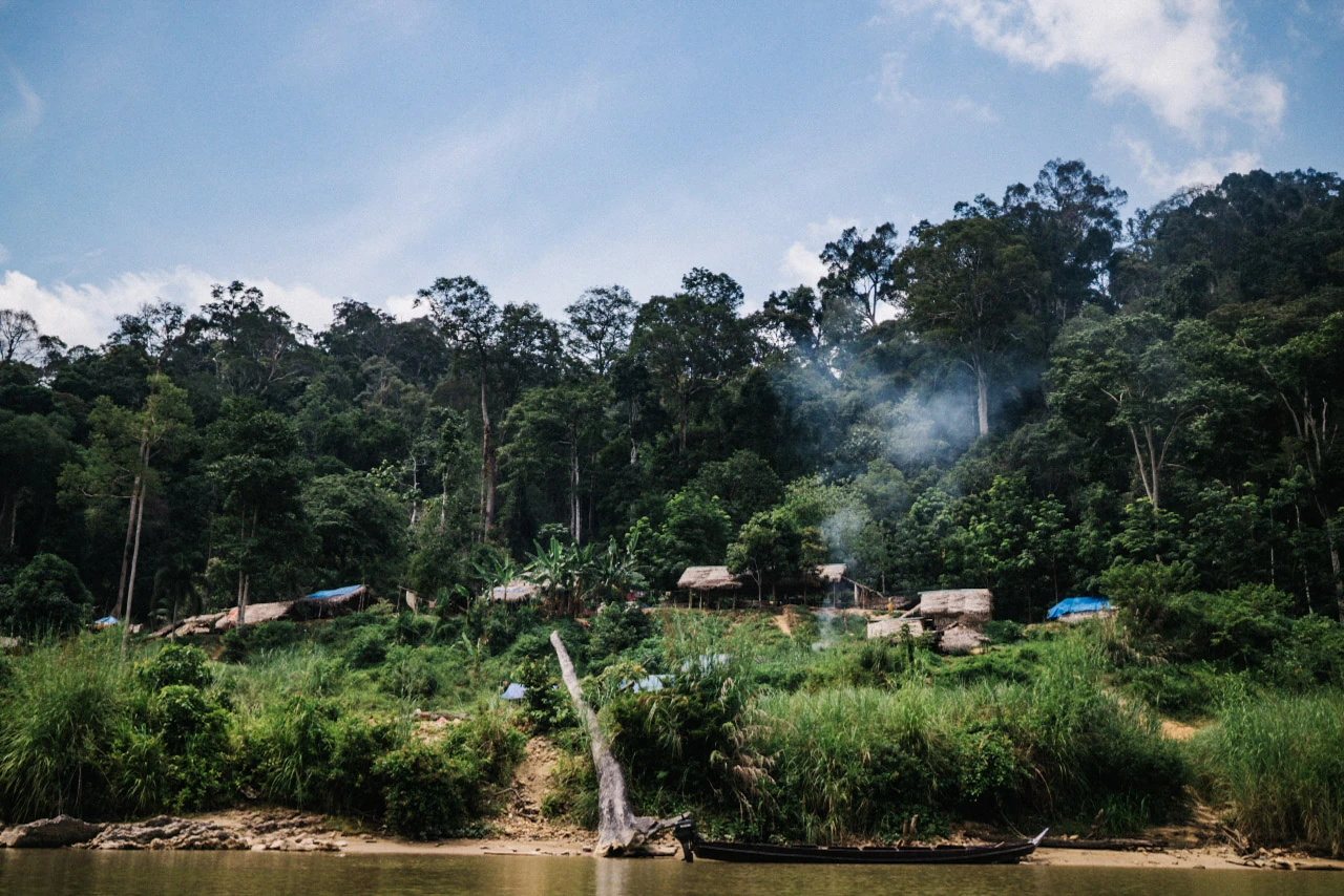 Pale smoke rises from a green riverbank with small buildings.