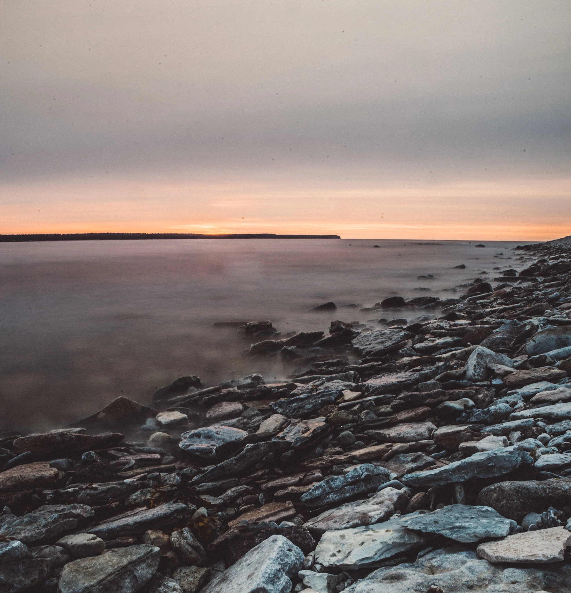 A beach of pale stones and softly rendered water beneath a narrow pink horizon.