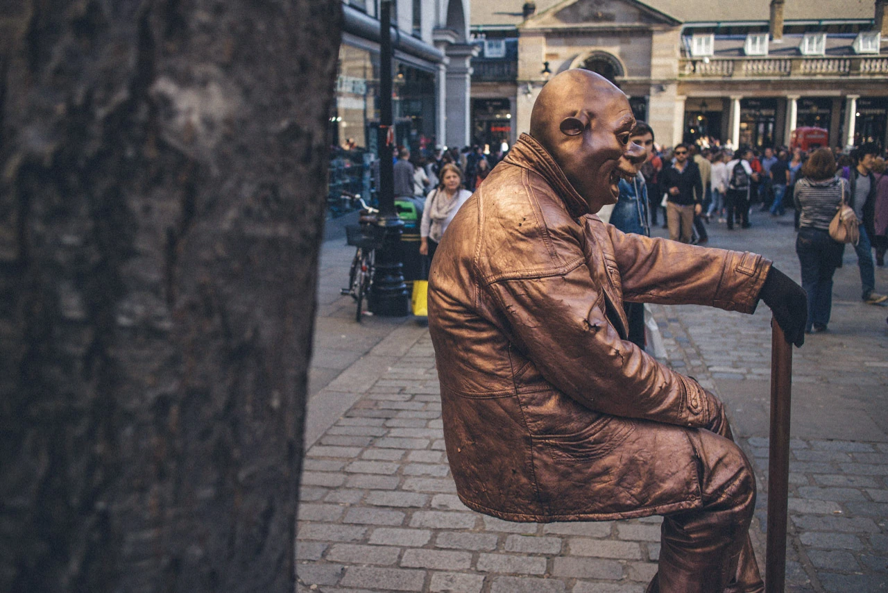 A bronze-painted street performer sits still in front of a busy street.