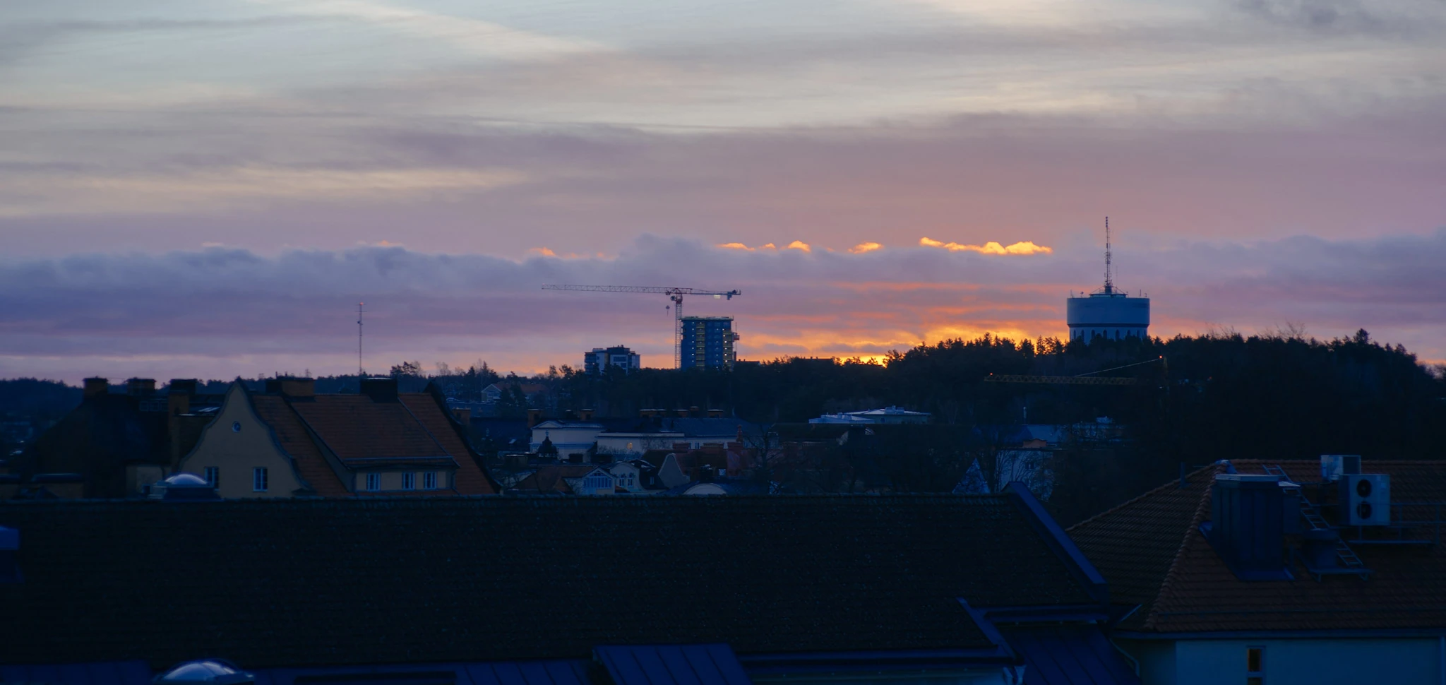 Dark roofs and distant towers beneath a narrow orange band in a blue evening sky.