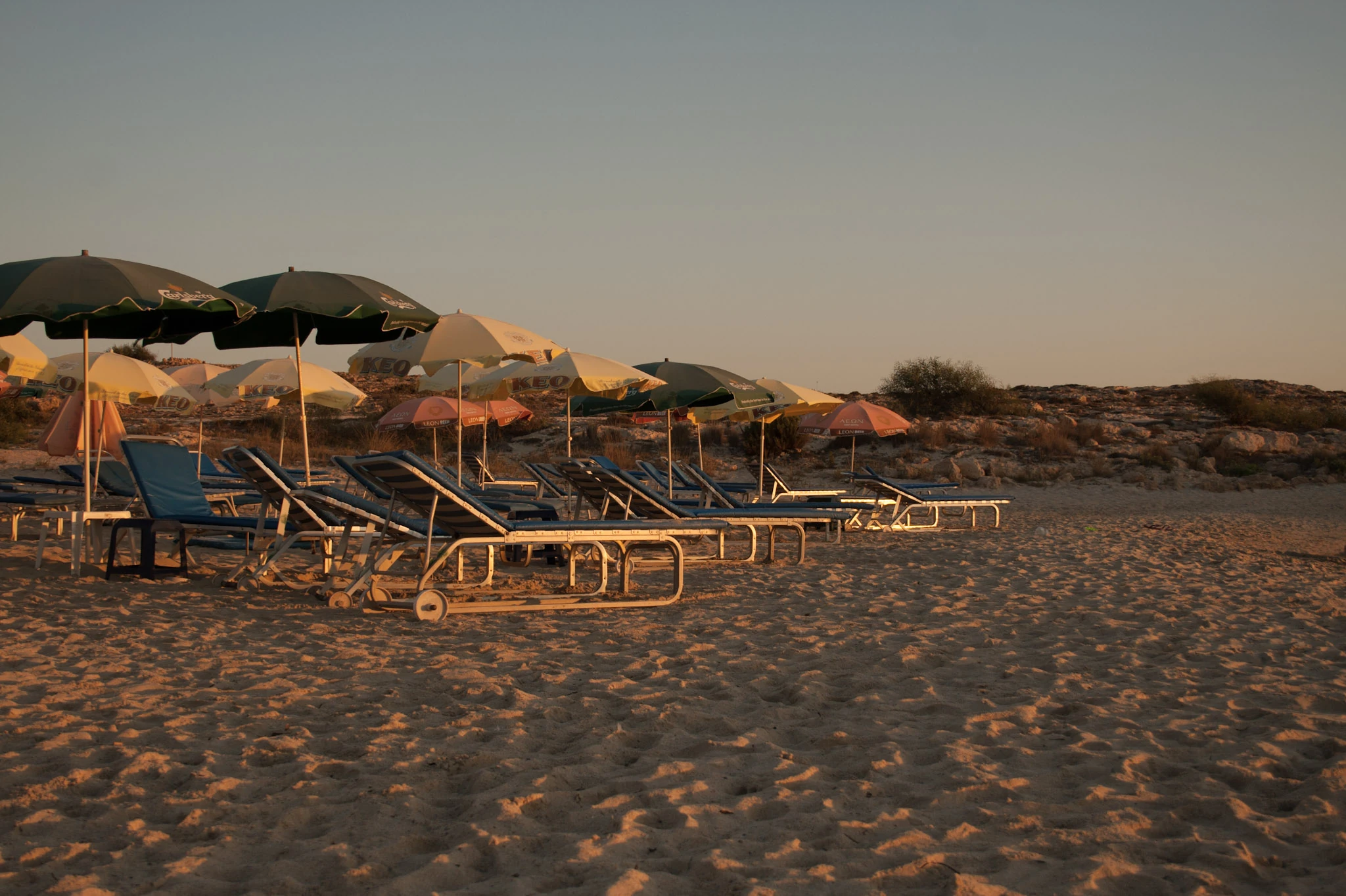 Empty loungers and parasols stand in warm evening light on a sandy beach.