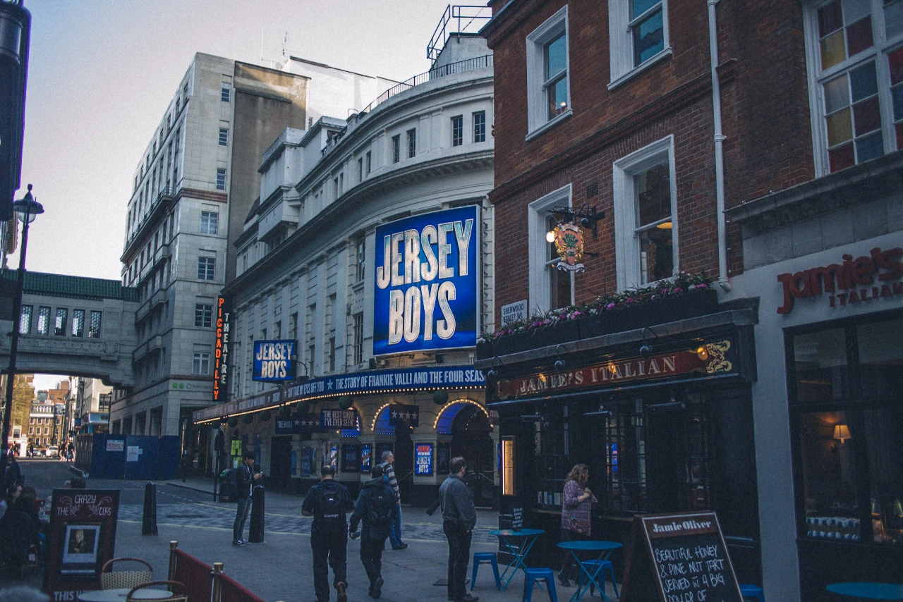 People on the pavement outside a London theatre with a large blue Jersey Boys sign.
