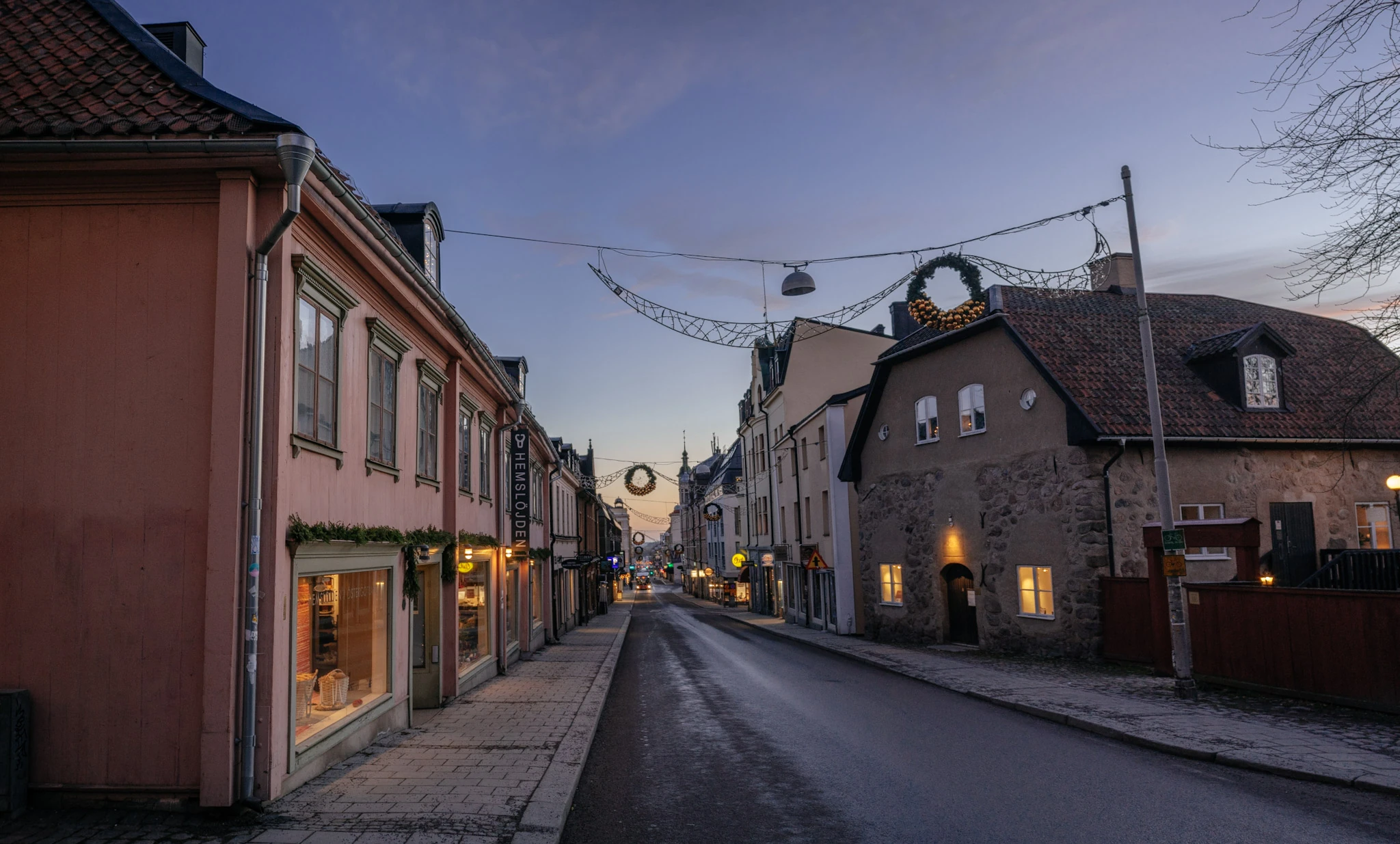 A nearly empty street between low buildings with lit windows beneath a pale evening sky.