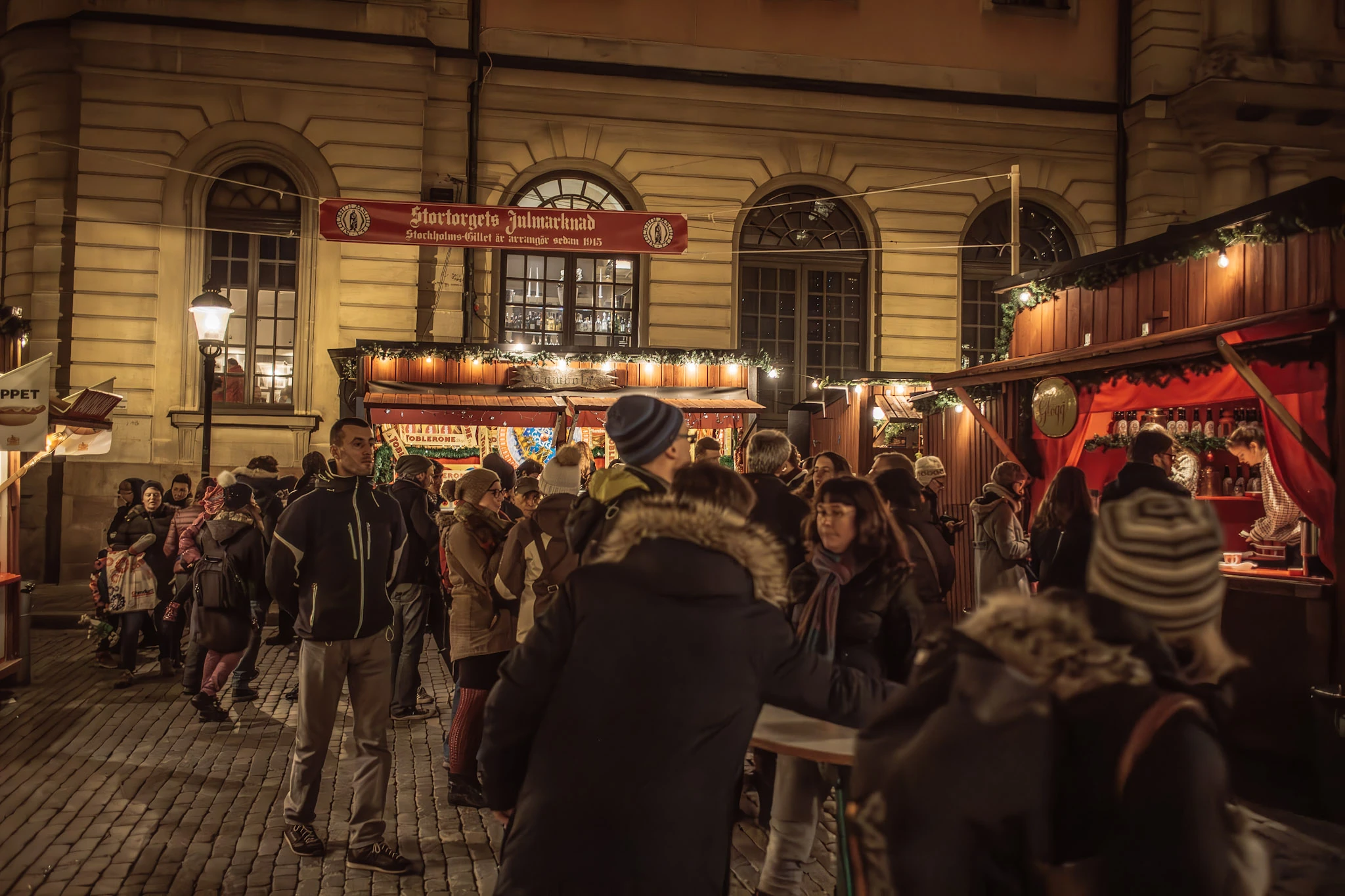 People between illuminated market stalls in front of a stone facade at night.