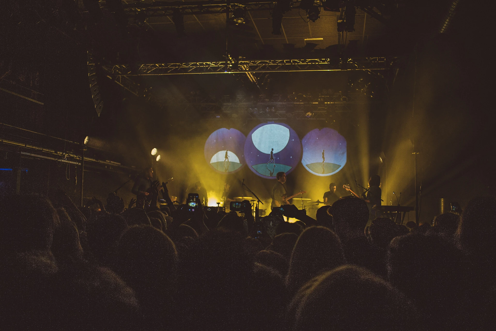 A band on a yellow-lit stage with three circular projections and an audience in front.