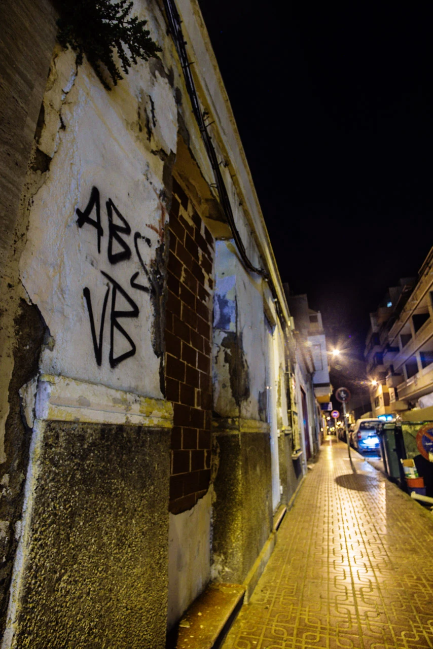A worn wall with graffiti beside a narrow illuminated alley at night.