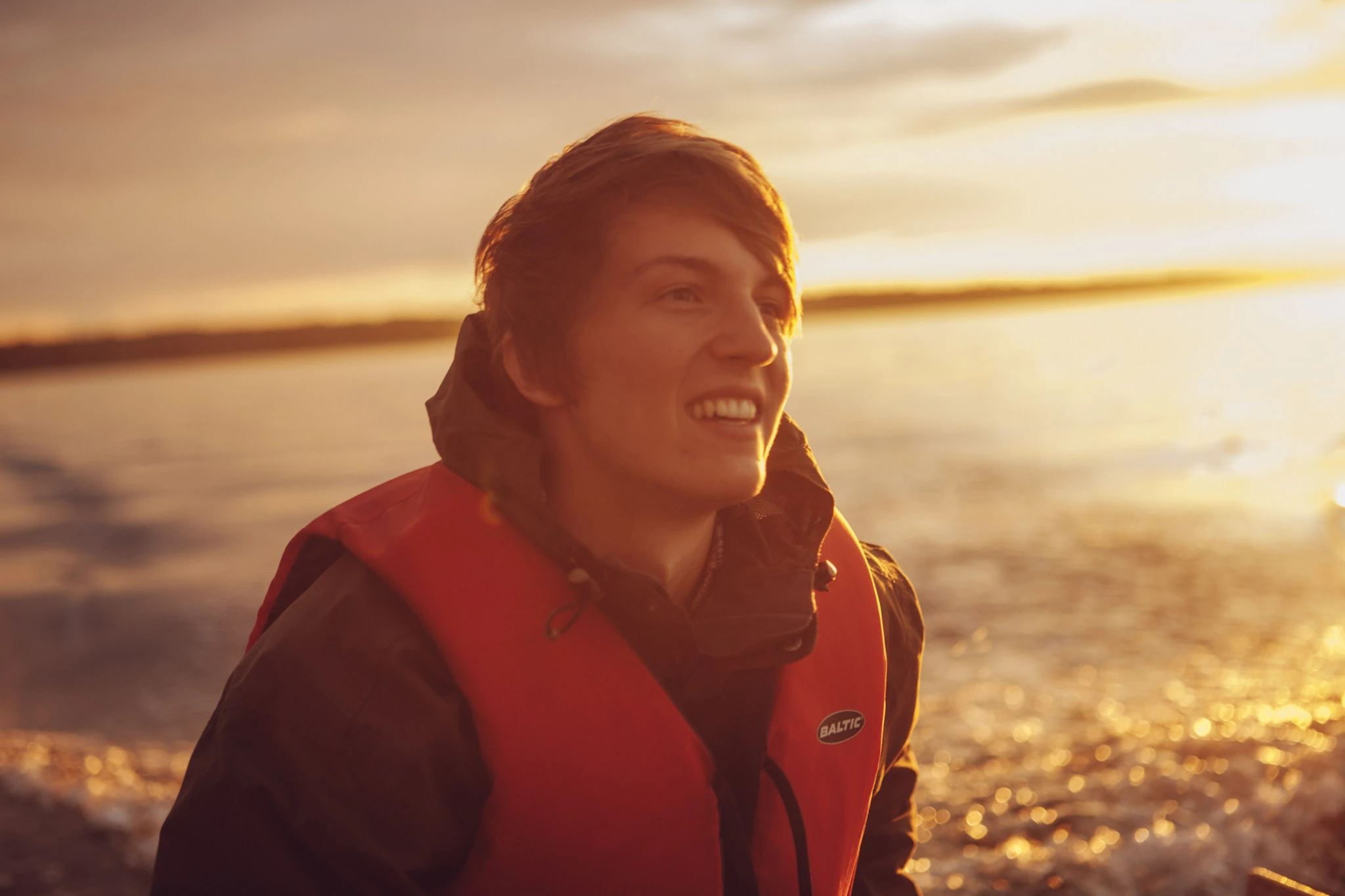 En ung man med röd flytväst skrattar i varmt kvällsljus vid havet.