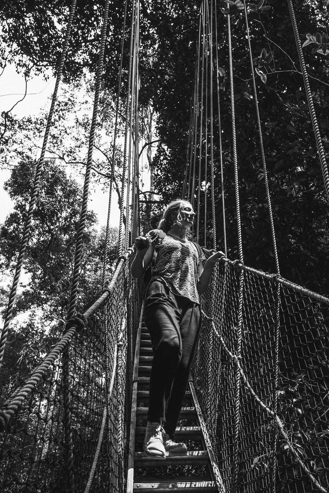 A person walks along a narrow suspension bridge between tall trees in a black-and-white photograph.