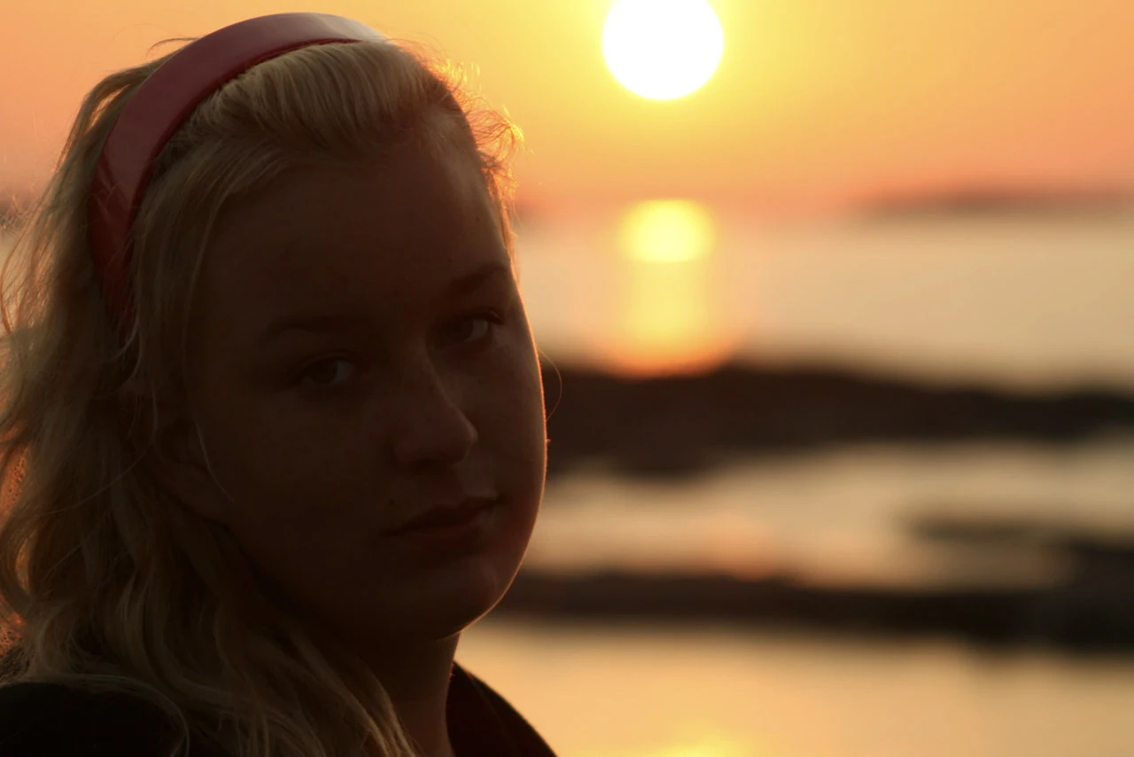 En blond kvinna porträtteras vid havet med solen bakom huvudet.