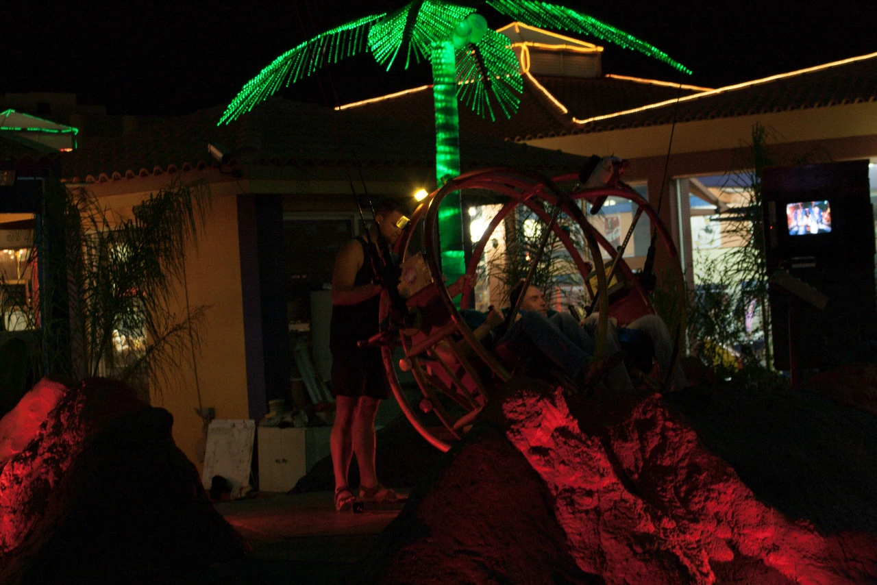 An artificial palm glows green above a large metal wheel in front of a building in red light and darkness.