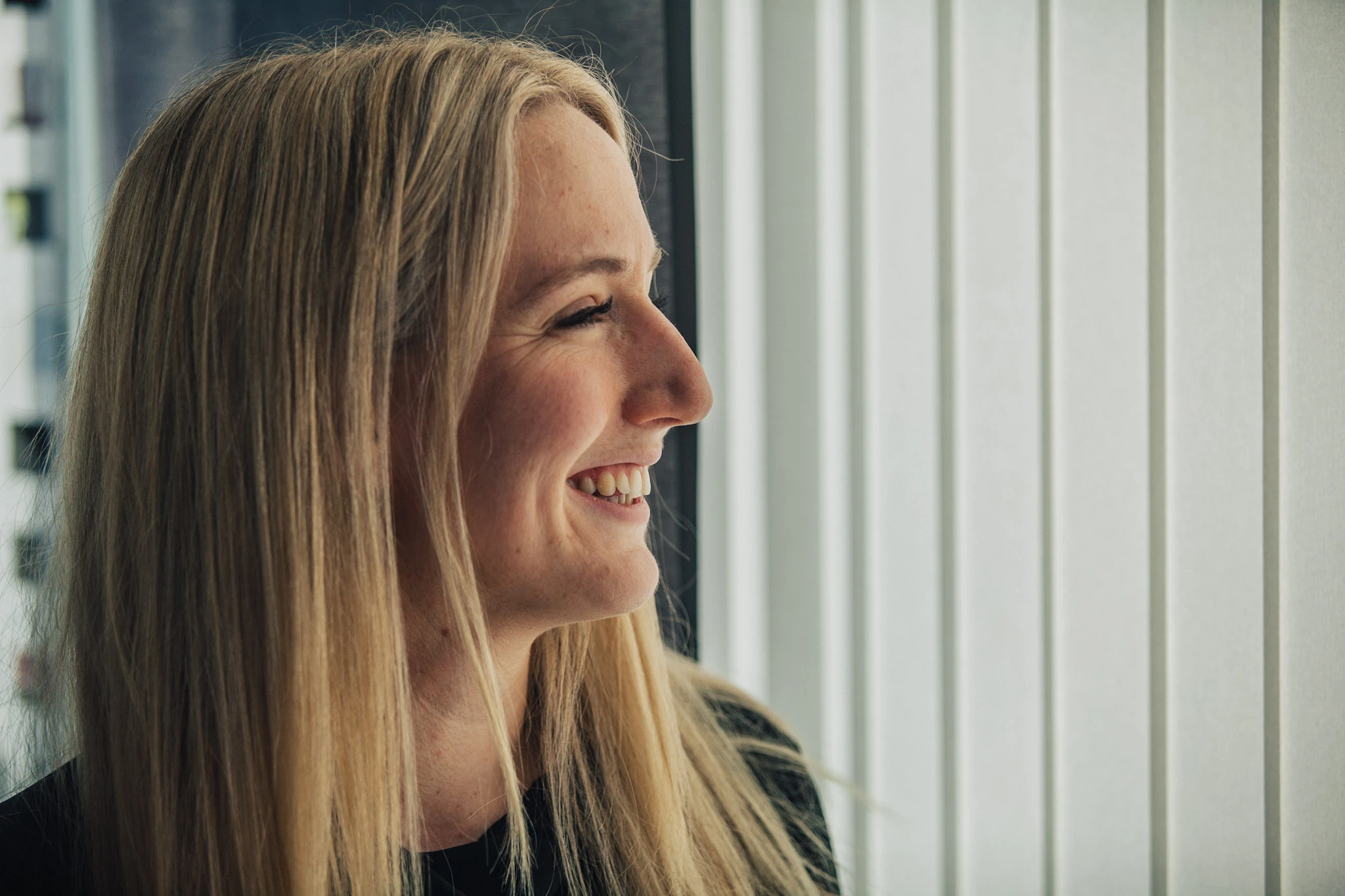 En blond kvinna ler i profil intill ett kontorsfönster.