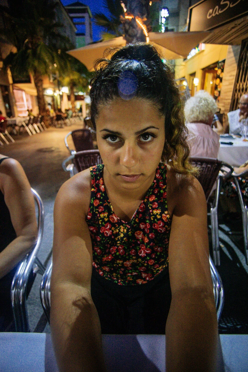 A woman leans forward and looks towards the camera with the night market behind her.