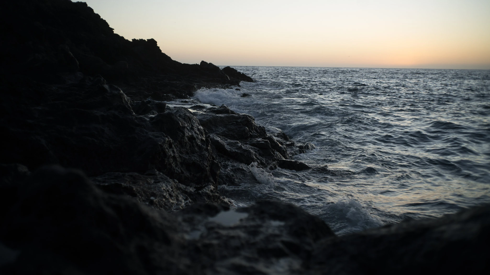 A dark rocky shore meets a rippling sea below a narrow warm horizon.