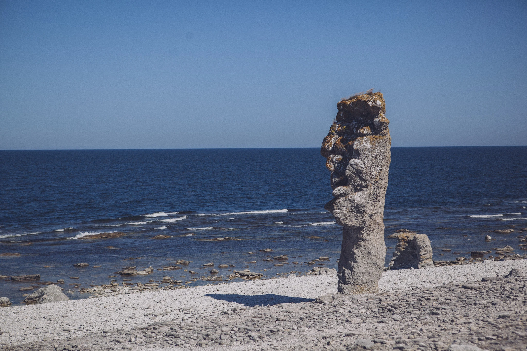 En ensam hög stenformation reser sig på en ljus strand vid blått hav.