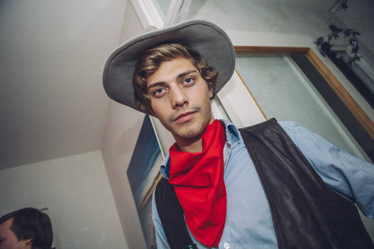 En ung man i cowboyhatt och röd scarf lutar sig mot kameran.