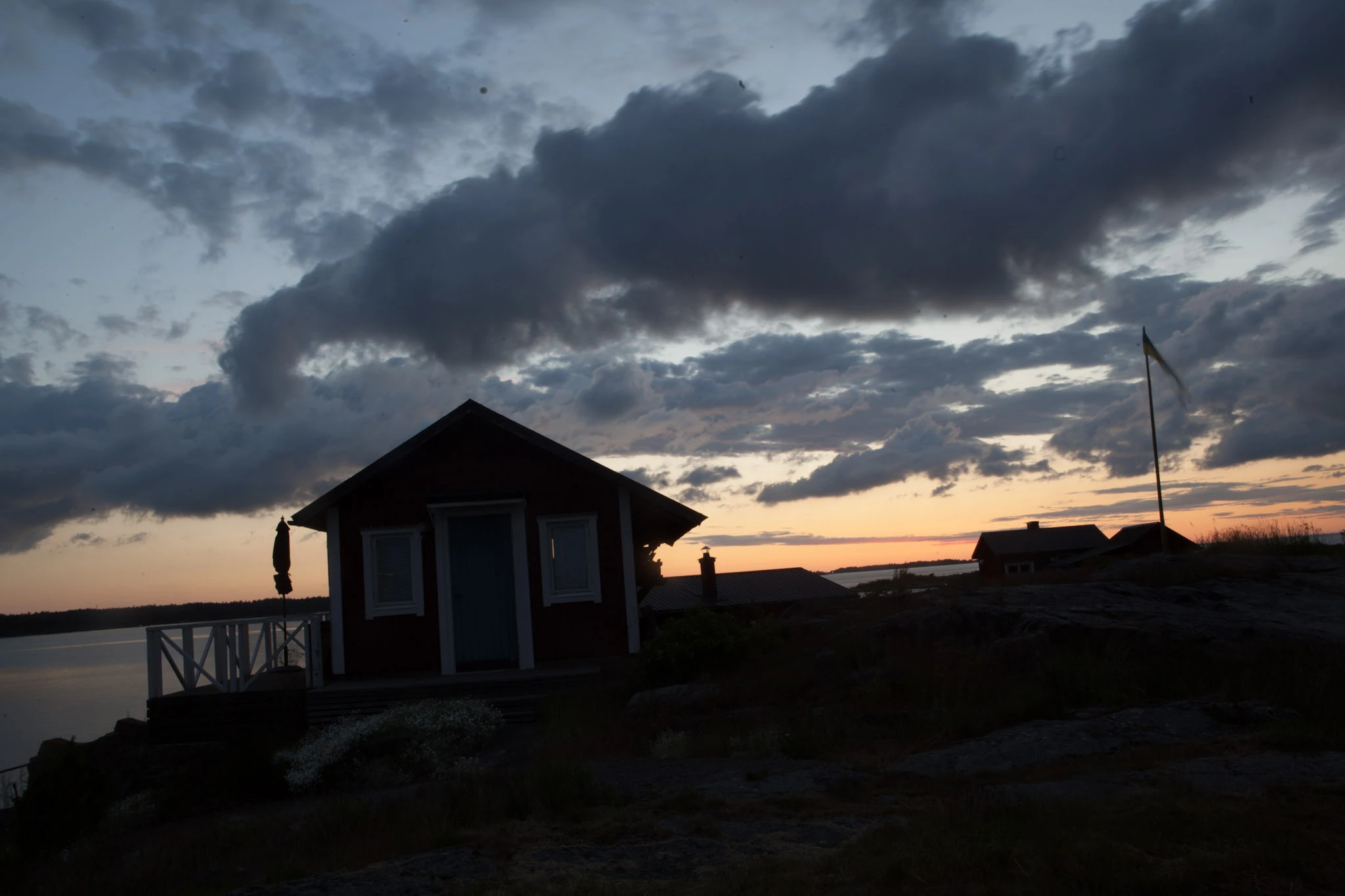 A small wooden building stands in silhouette by water beneath dark clouds and a warm horizon.