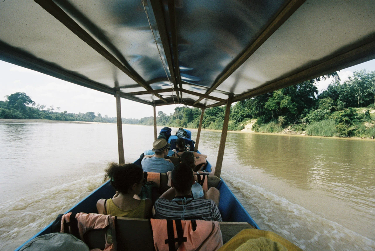 Passengers sit beneath the roof of a narrow boat on a brown river.