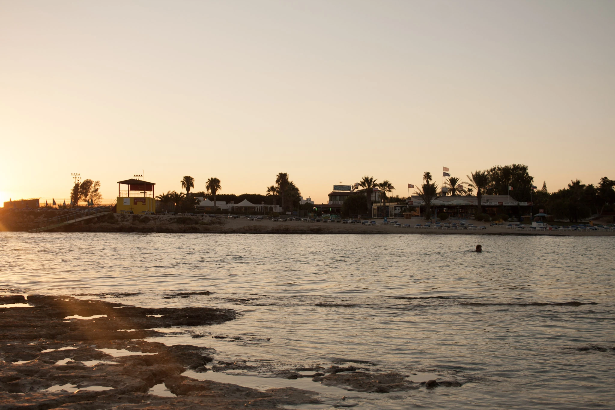 A low sun shines over water towards palms and small buildings on the opposite shore.