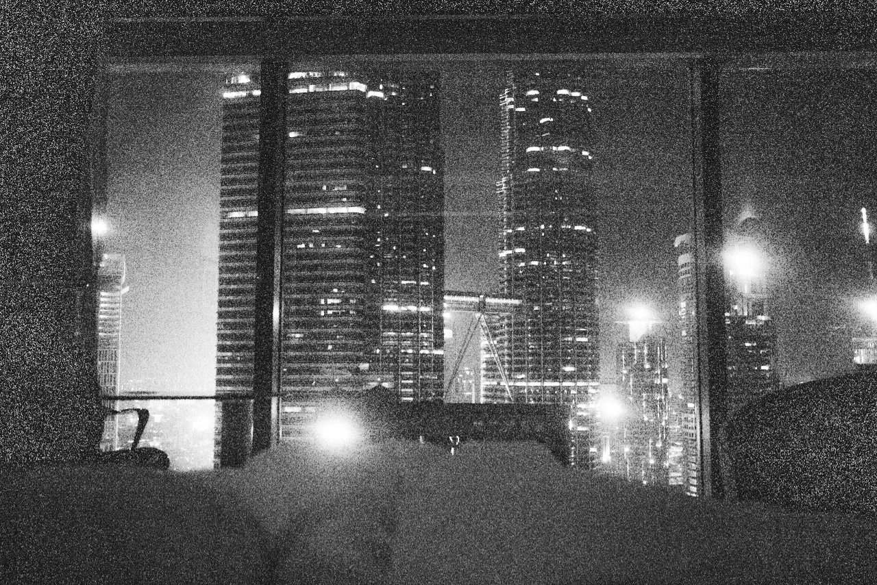 Tall city buildings are seen through a dark window in black and white.