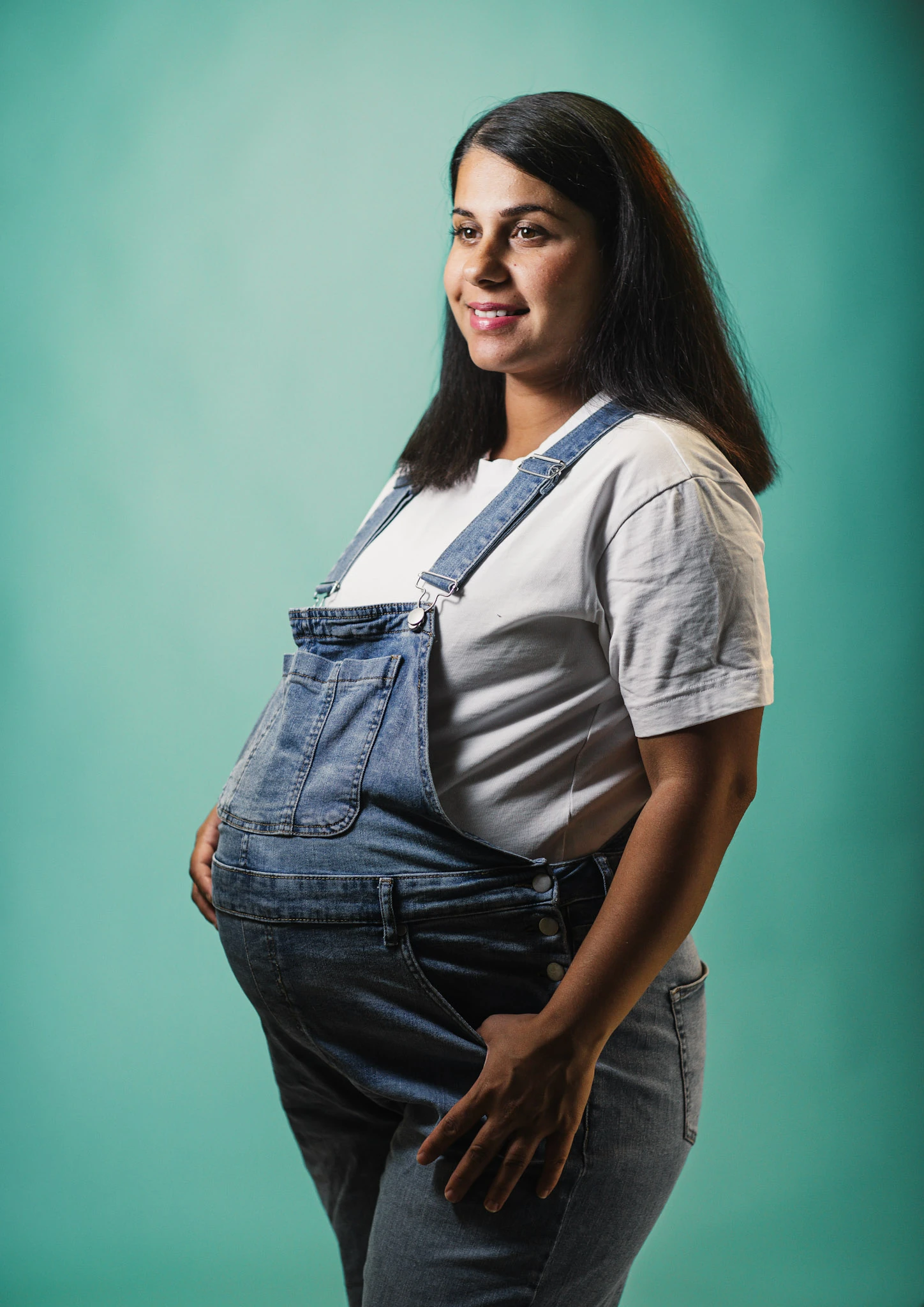 En gravid kvinna i blå hängselbyxor mot grönblå studiofond.