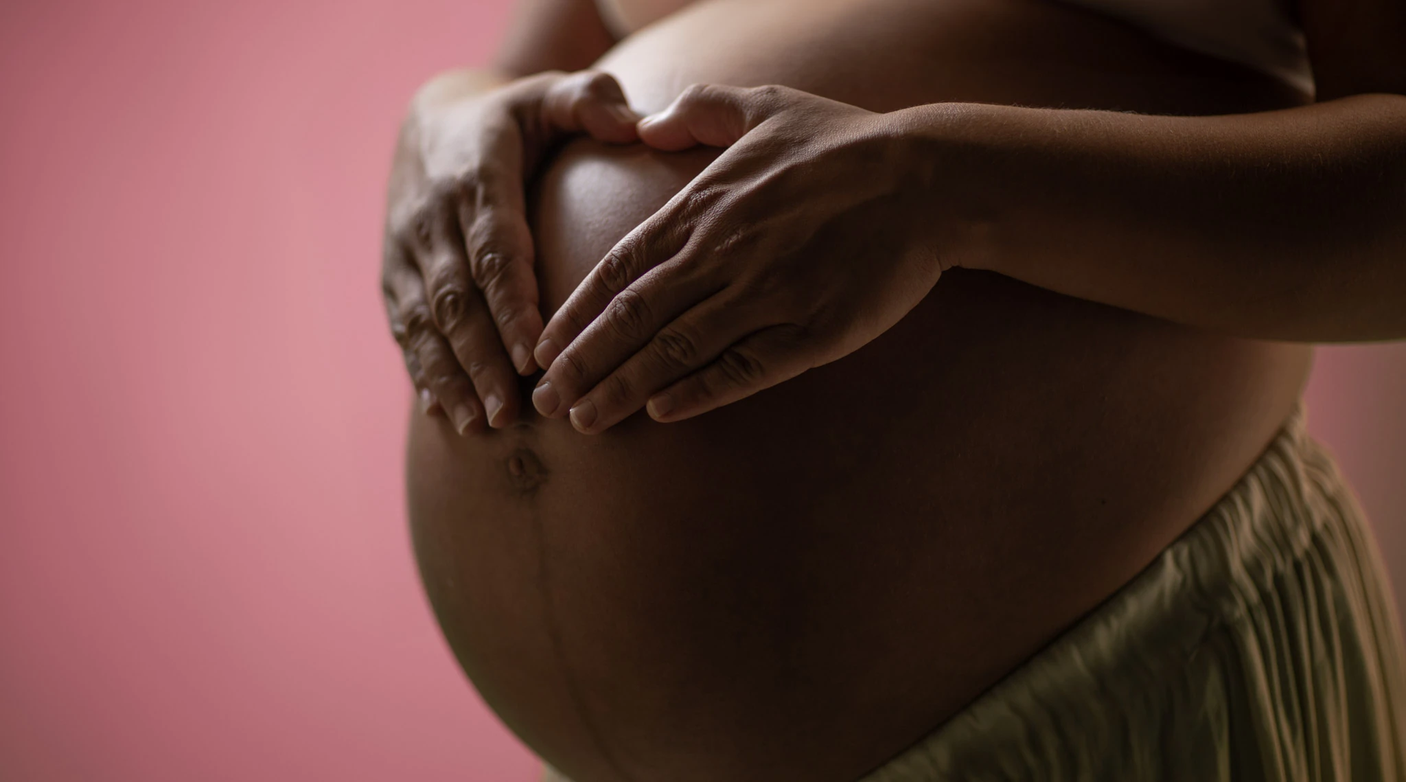 Händer vilar på en gravid mage mot en rosa bakgrund.
