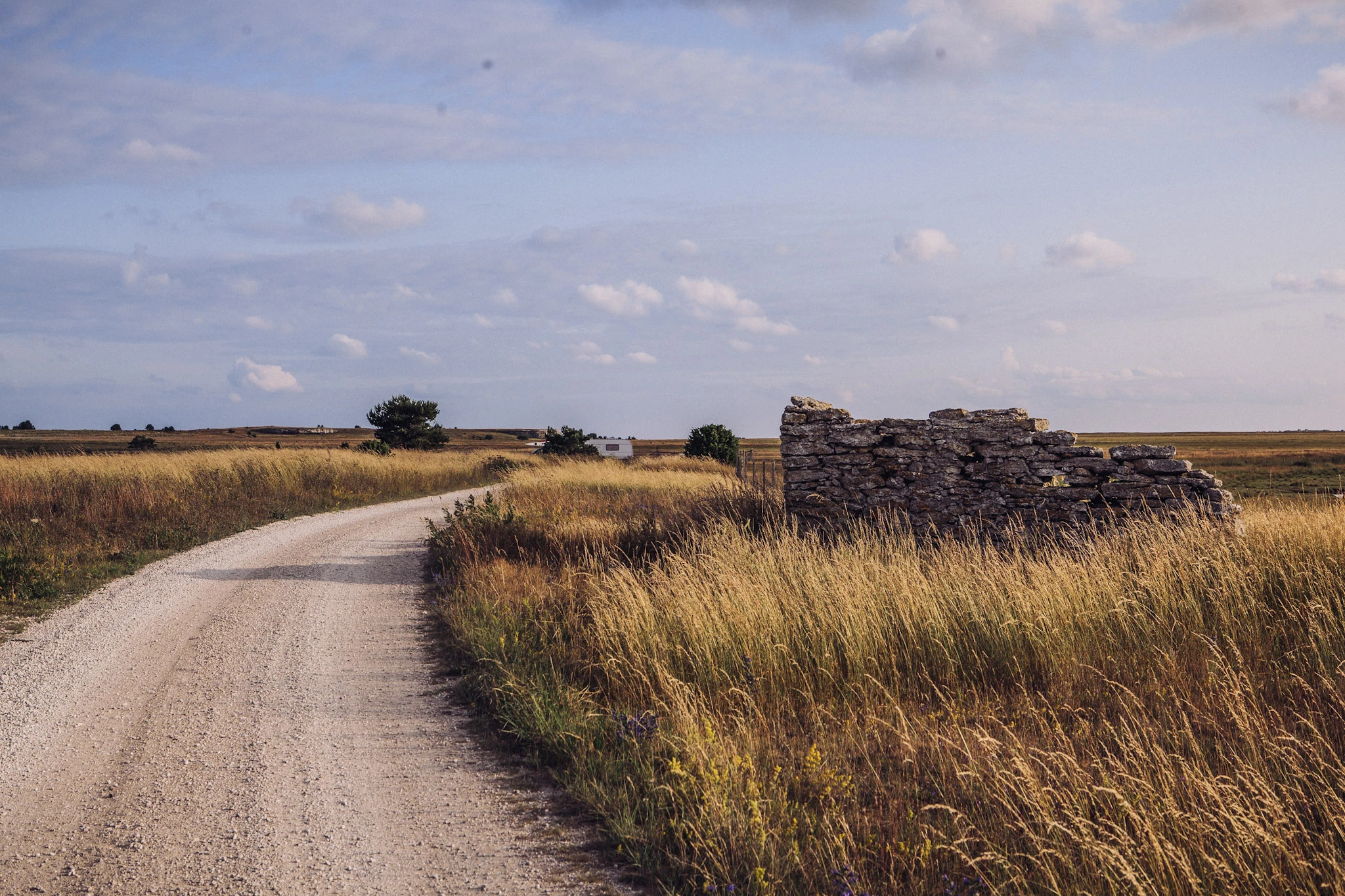 A gravel road winds through dry grass past a low stone wall.