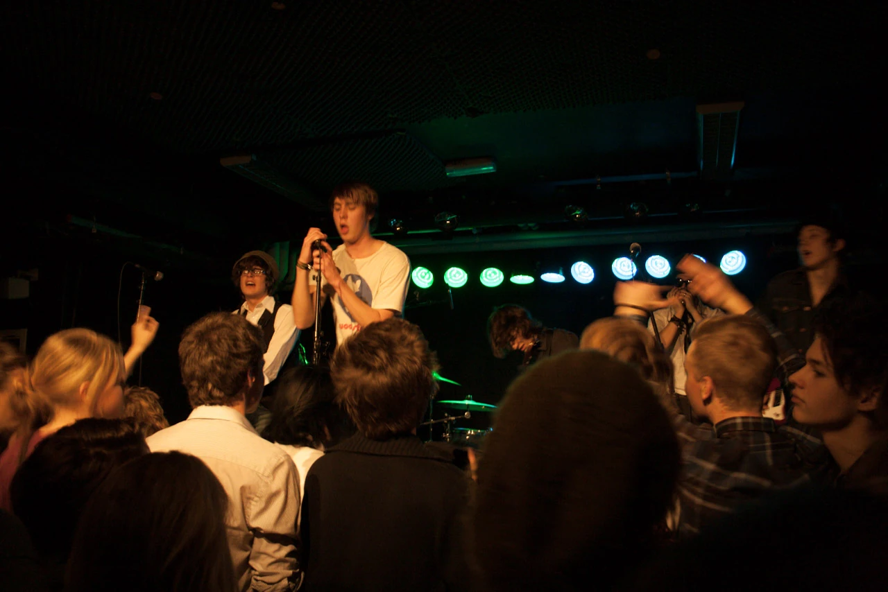 A singer in a white shirt surrounded by a band and an audience close to the stage.