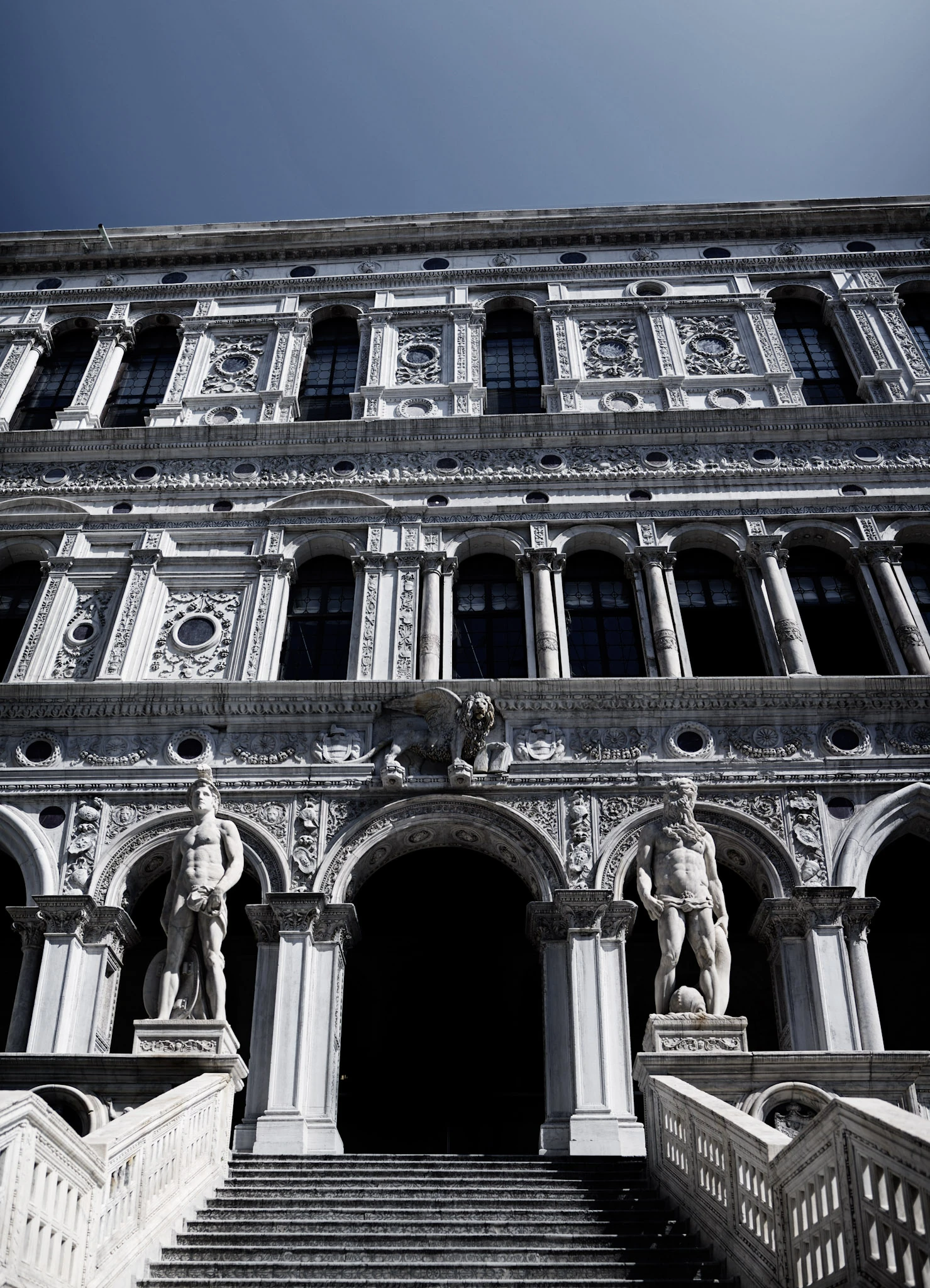 A black-and-white view of a broad stone staircase with sculptures in front of a building with large arched openings.