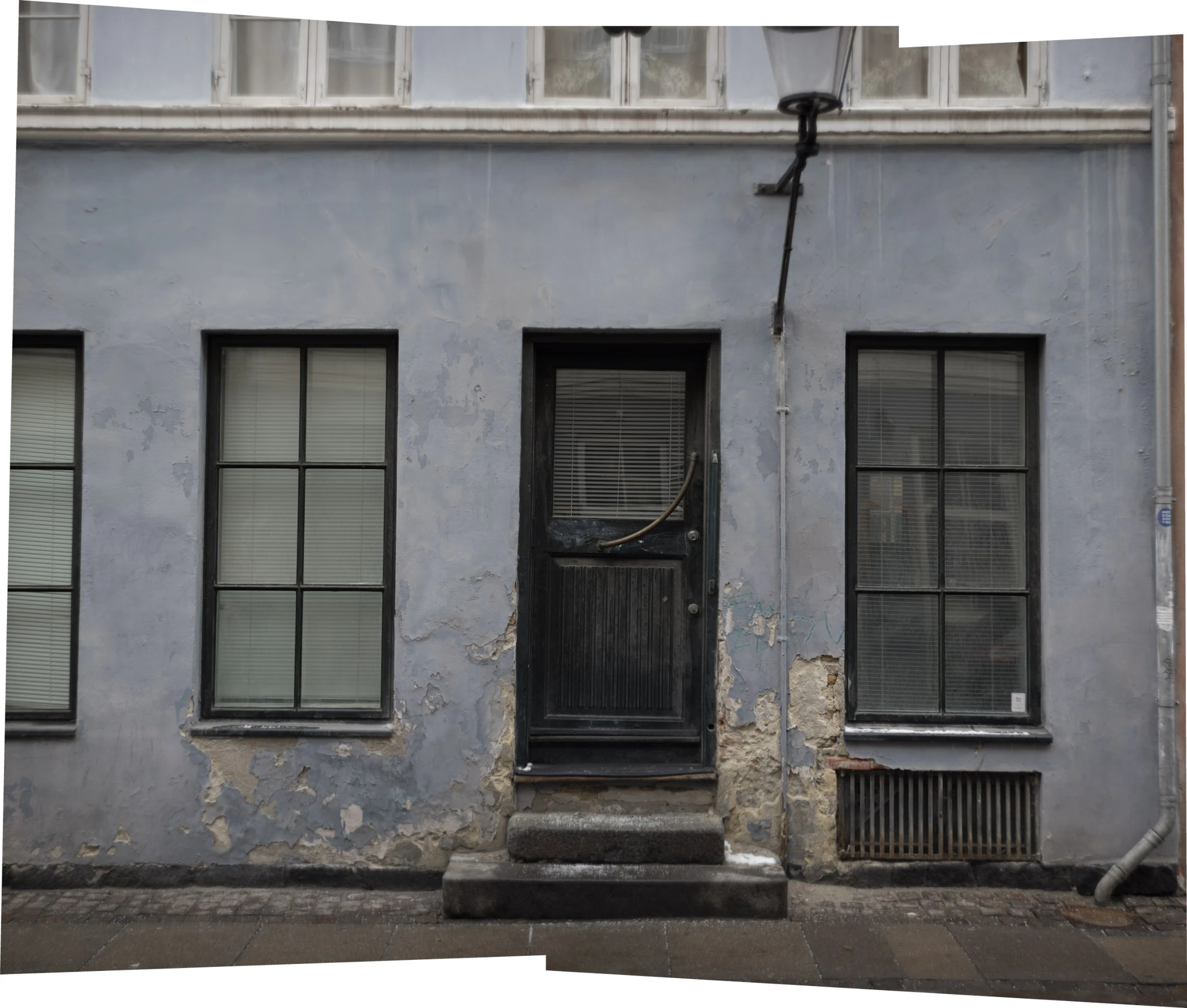 A grey facade with two tall windows, a narrow dark door and a drainpipe.