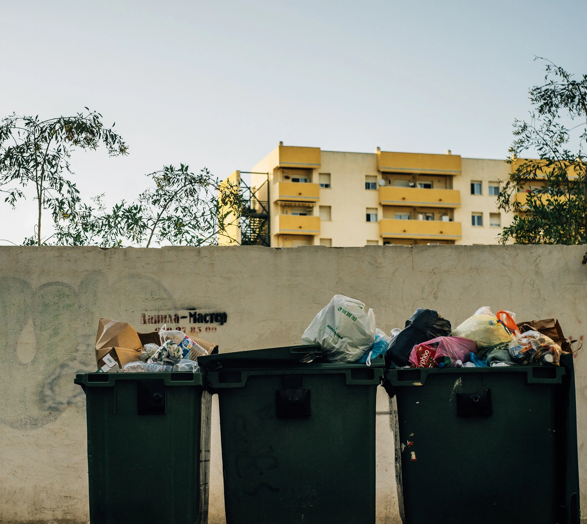 Three green bins filled with bags and cardboard in front of a wall and a yellow apartment building.