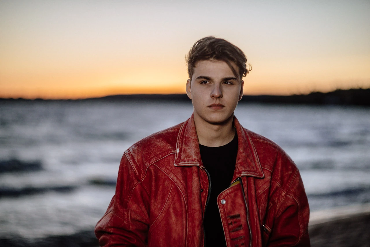 A young man in a red leather jacket stands before water and an orange twilight sky.