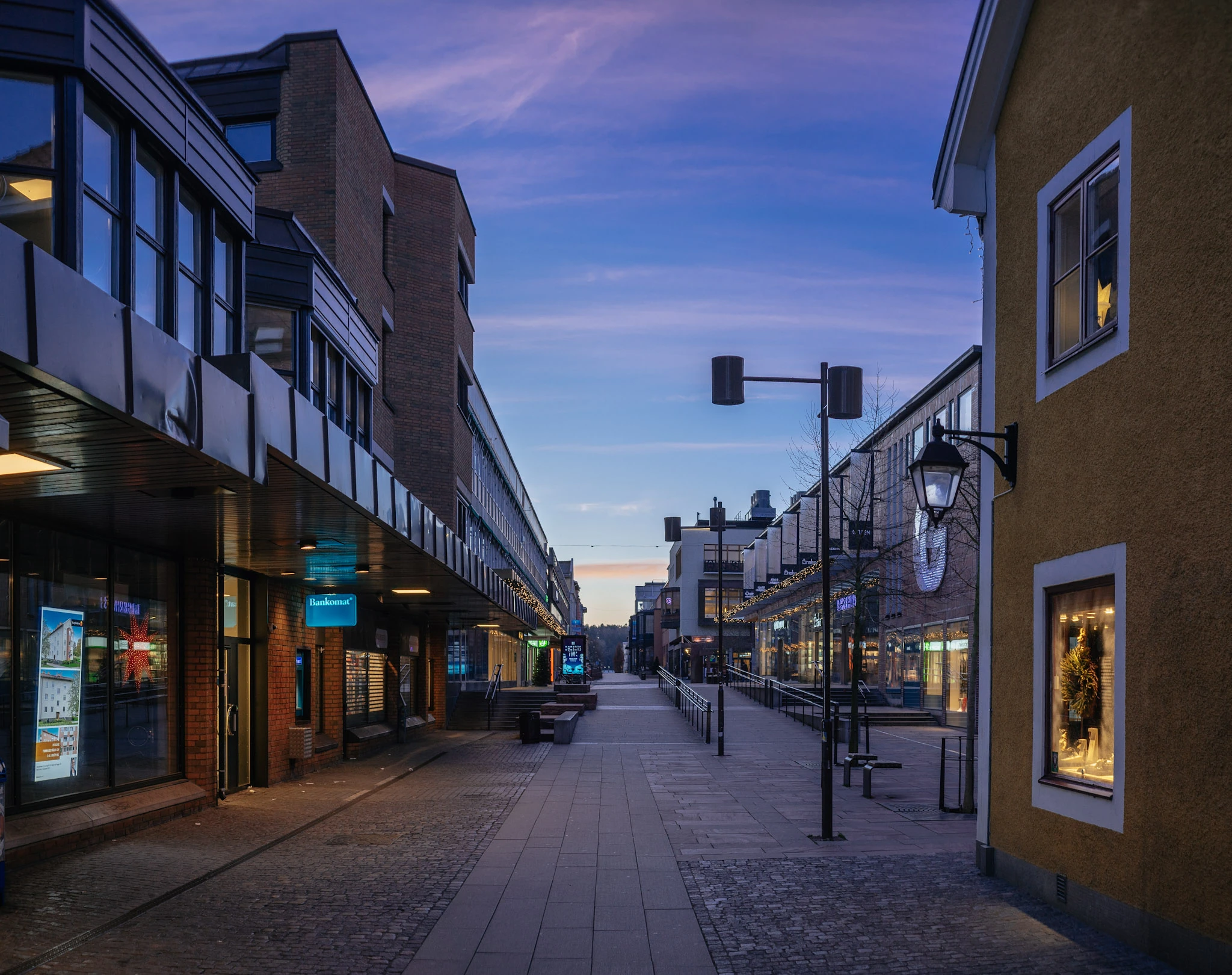 A straight city street between dark facades and a yellow building beneath a blue twilight sky.